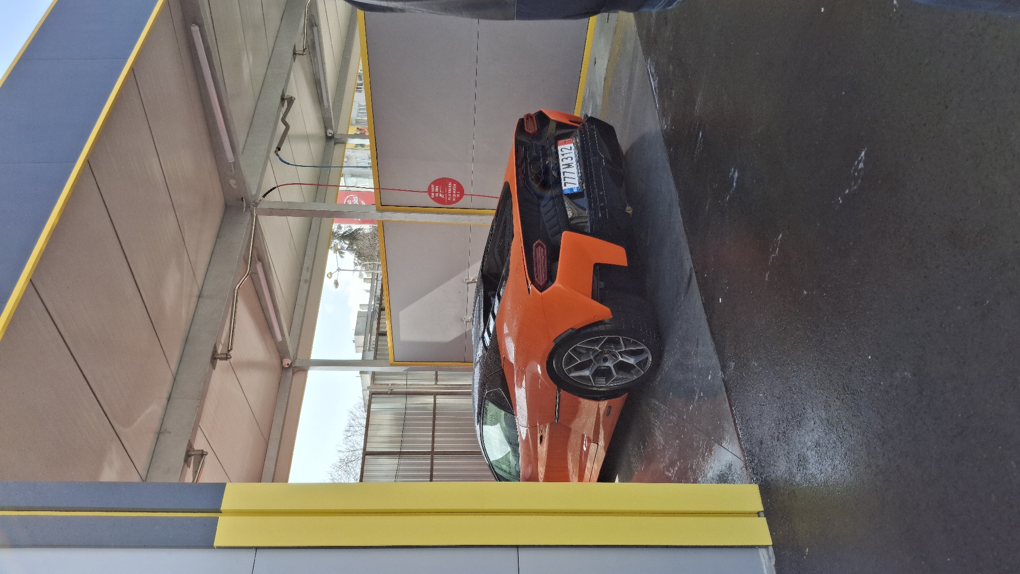 Here is a forensic description of the image:
An orange Lamborghini sports car is positioned within a car wash facility. The vehicle's license plate is partially visible, displaying "777 M 312". The car's surface appears to be wet, indicating it has recently been washed or is in the process of being washed. The car wash facility features a metal and concrete structure with visible lighting fixtures and overhead pipes. A red sign is posted on the wall. The floor of the car wash is wet, and the area surrounding the car suggests it's a drive-through facility. The city location is not identifiable from the image.