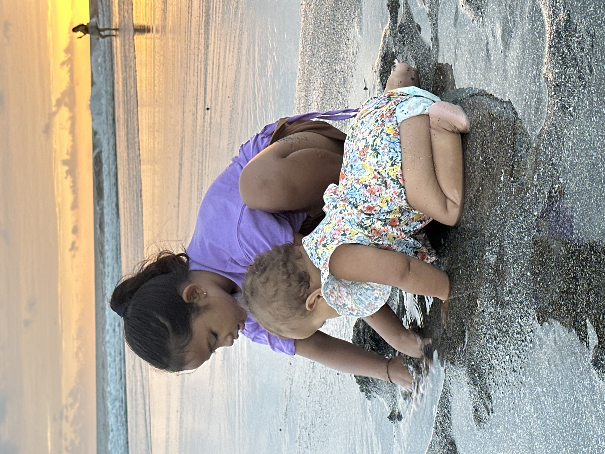 Two young girls are playing in shallow water on a beach at sunset. The older girl, wearing a purple t-shirt, is crouched down and looking at the water. The younger girl, in a floral-print dress, is also crouched and playing with the water. In the background, the ocean meets the sky, with waves and a person standing in the water. The sand is dark and wet, reflecting the warm colors of the sunset. The location is a beach in North Kuta, Indonesia.