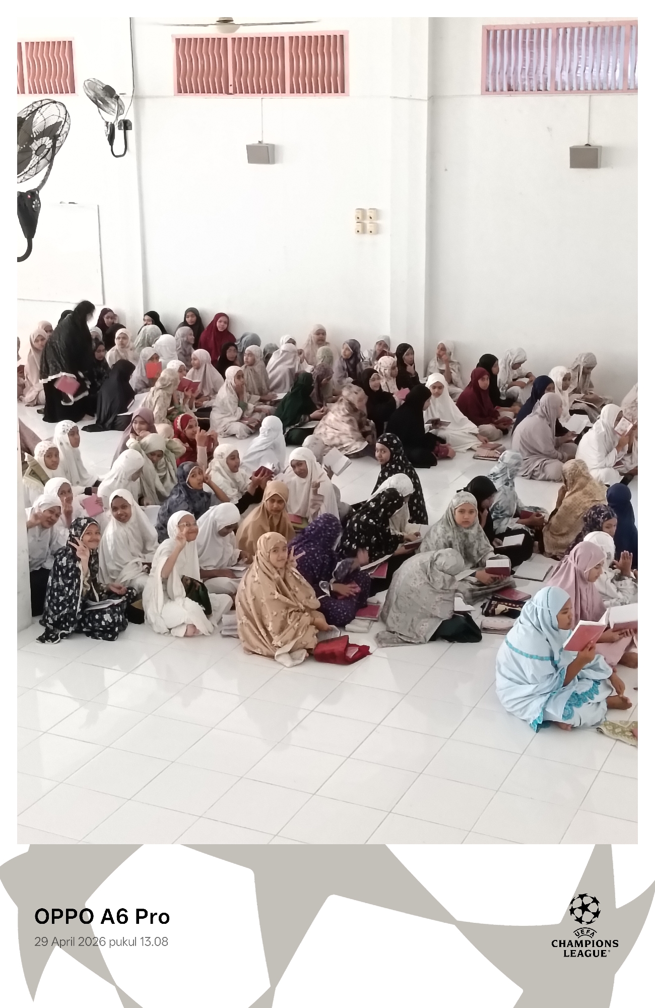 A large group of approximately 50-60 young girls and some adults, predominantly wearing hijabs and modest clothing in various colors, are gathered indoors in what appears to be a religious school or educational setting in Lhokseumawe, Indonesia. They are seated on a white tiled floor in a spacious room with plain white walls. High on the walls, there are two pink louvered vents and wall-mounted speakers. Two ceiling fans and an electrical outlet are also visible.
The individuals are engaged in a study or reading session, with many holding open books, likely religious texts, and appearing to read or recite from them. Several of the girls are looking towards the camera, with many smiling cheerfully and some making peace signs, indicating a relaxed and positive atmosphere. A few adults are also present, seemingly supervising the session.
The photo was captured on "29 April 2026 pukul 13.08" (1:08 PM) using an "OPPO A6 Pro" phone, as indicated by the watermark in the bottom left corner. Another watermark in the bottom right corner displays the "UEFA CHAMPIONS LEAGUE" logo and text. The indoor setting and the presence of fans suggest it might be a warm climate. The attire and activity strongly indicate a cultural and religious context common in Indonesia.