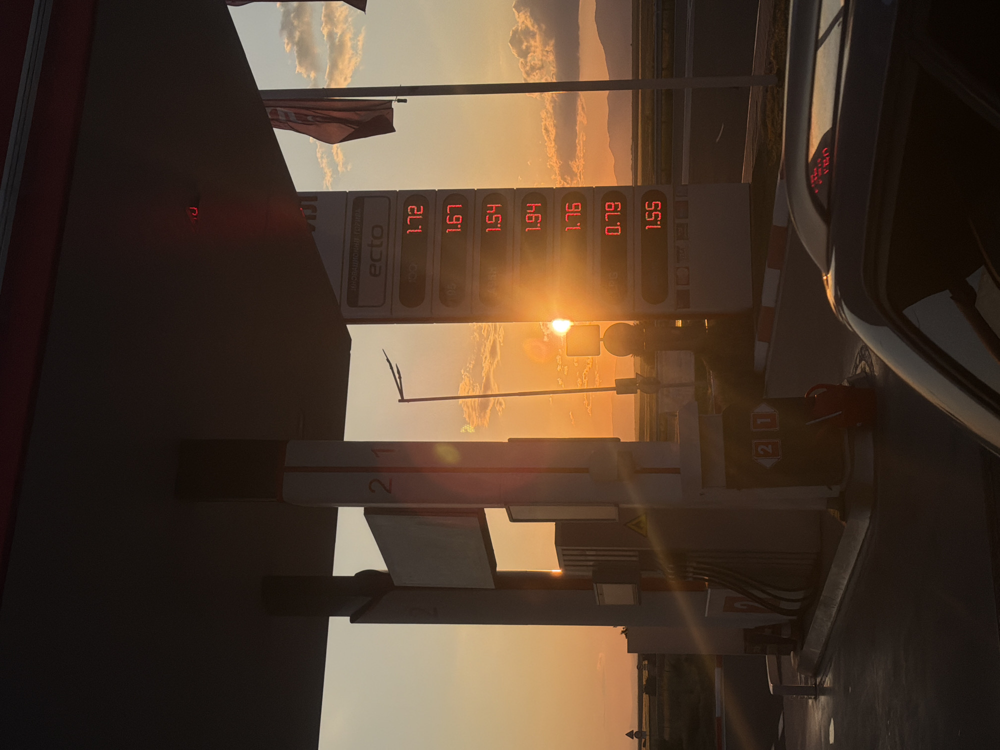 The image shows a gas station with a sign displaying fuel prices: 1.72, 1.67, 1.54, 1.94, 1.76, 0.79, and 1.55. The gas station is branded with "ecto" and the words "ИНОВАЦИОННИ ГОРИВА." A gas pump is visible, labeled with the numbers "2" and "1." A car is partially visible on the right side of the frame. The background shows a landscape with a setting or rising sun, casting a strong light and creating silhouettes. The location is Vodenichane, Bulgaria.