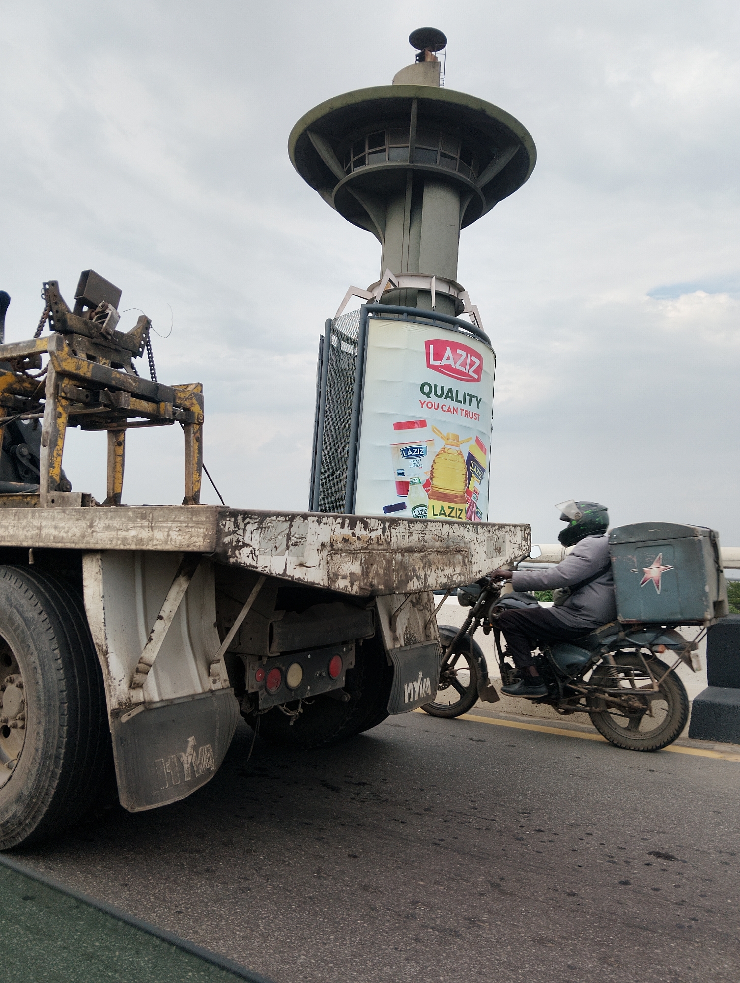 The image shows a flatbed truck carrying a large cylindrical advertising billboard. The billboard displays the brand "LAZIZ" with the slogan "QUALITY YOU CAN TRUST" and images of their products. To the right of the truck, a person wearing a grey jacket and a helmet is riding a motorcycle. The motorcycle has a large cargo box mounted on the back. The scene appears to be on a road in Alagbede, Nigeria, with a concrete barrier and a yellow line visible on the asphalt. The sky is overcast. The truck has a yellow and black metal structure on its front, likely a loading mechanism. The billboard is attached to a tall concrete structure resembling a water tower or a similar utility tower.