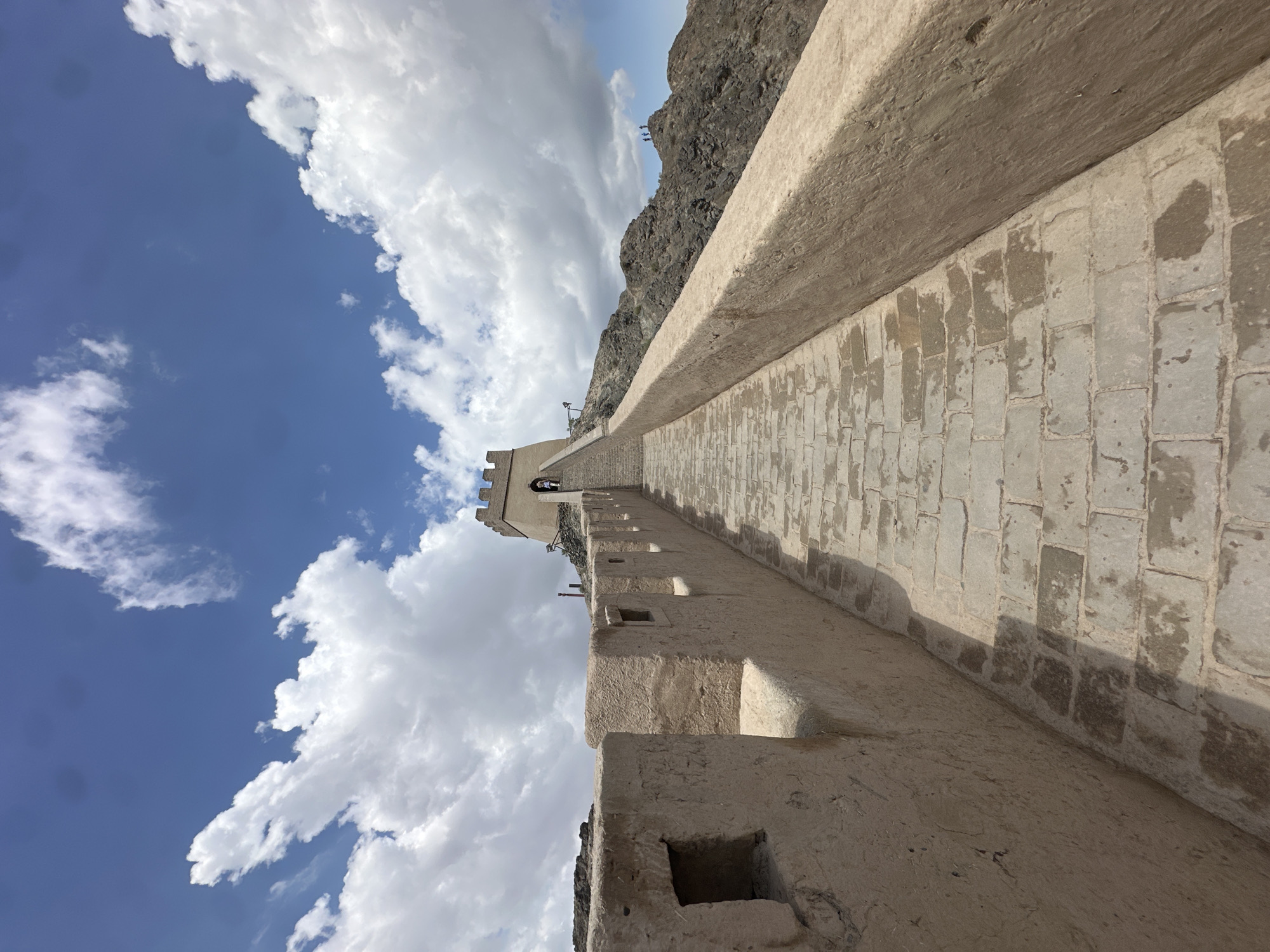 The image captures a section of the Great Wall of China near Jiayuguan, China. The wall, constructed from tan-colored bricks and stone, ascends a rocky, mountainous terrain under a bright blue sky dotted with fluffy white clouds. The perspective is looking up a stone-paved walkway that follows the contours of the wall, with crenellated battlements lining the left side.
A watchtower stands at the crest of the incline, its stone structure visible against the sky. Several individuals are present on the walkway and near the watchtower, appearing to be tourists. In the distance, on a rocky outcrop to the right of the wall, a small group of people can be seen. The environment is dry and arid, characterized by the rocky, rugged landscape. The lighting suggests it is daytime, with distinct shadows cast by the wall and its features on the walkway.