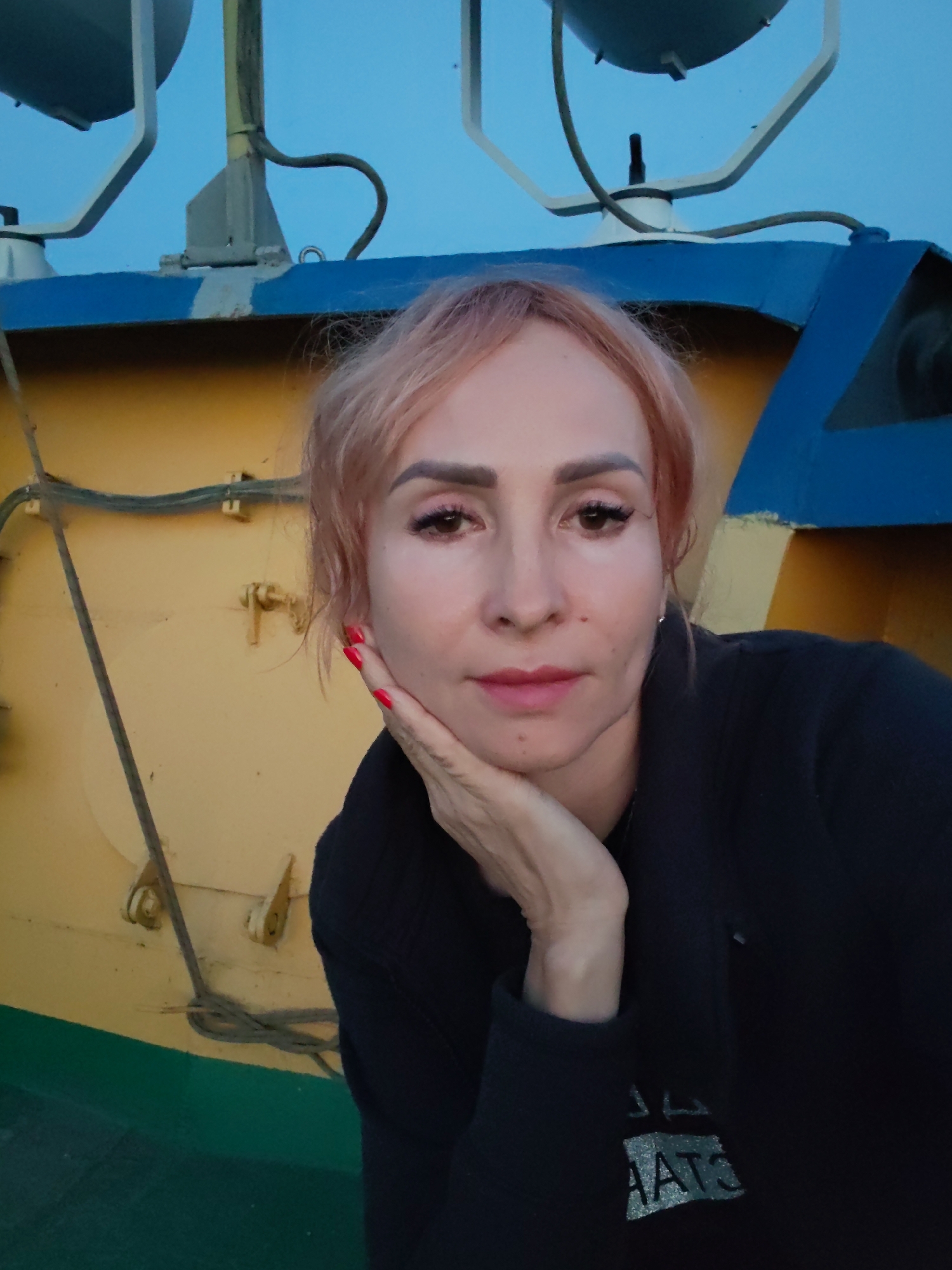 A woman with reddish-blonde hair and visible makeup is posing for a photo. Her right hand is resting on her chin, and her left side is cut off by the frame. She is wearing a dark jacket with a reflective, textured design on the left side of the chest. The background consists of a yellow, blue, and green structure, possibly part of a boat or industrial equipment, against a clear blue sky. The scene appears to be outdoors, likely in the city of Bol'shie Mkhi, Russia, given the context.