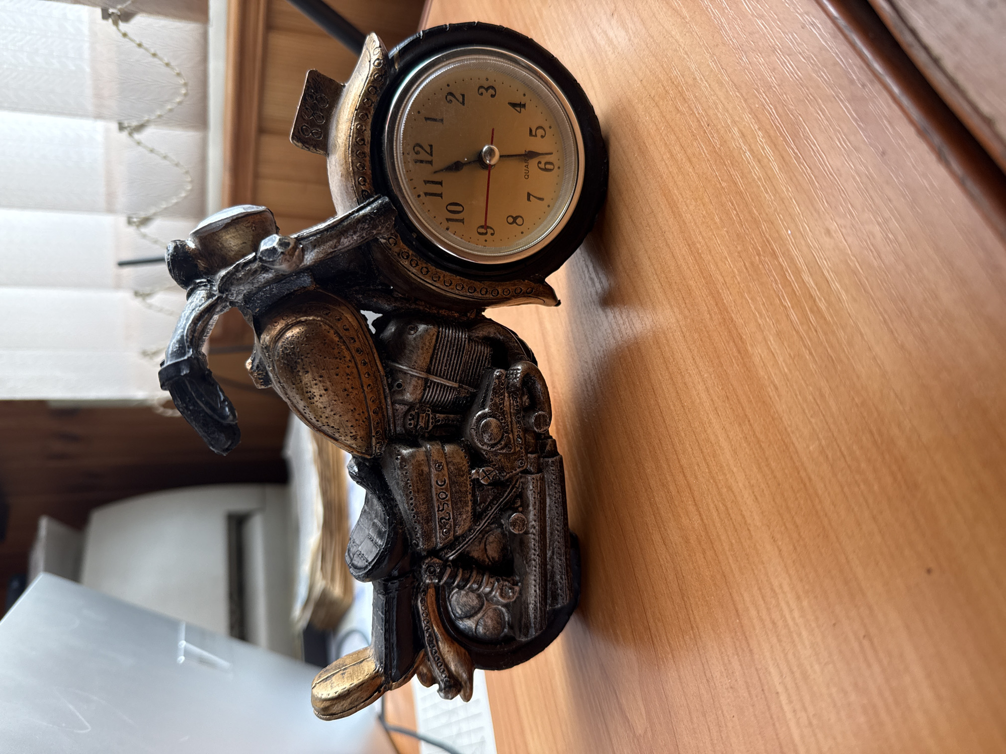 A close-up, eye-level, medium shot captures a decorative clock in the shape of a motorcycle, placed on a wooden desk. The motorcycle clock is detailed, with a bronze and black finish, and a clock face incorporated into the front wheel. The clock hands indicate the time is approximately 12:07. The background is blurred, showing a white printer-like object and sheer curtains. The lighting is natural, coming from the right, casting a soft shadow on the desk. The image appears to be taken indoors, in a well-lit room.