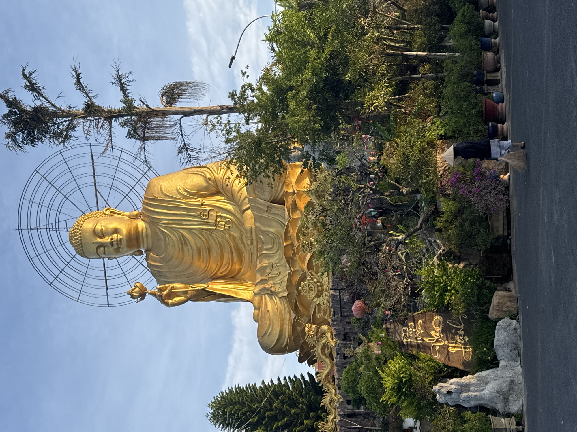 A golden Buddha statue stands in a landscaped garden in Lâm Viên, Đà Lạt, Vietnam. The statue is a large, golden figure with a halo-like structure behind the head, holding a lotus flower. Trees and foliage surround the base, including a decorative rock with gilded writing. A person wearing traditional conical hat sweeps along a paved path near the statue. Vases or plant pots are lined up along the paved path, suggesting a well-maintained area. The sky is light blue with wispy clouds.