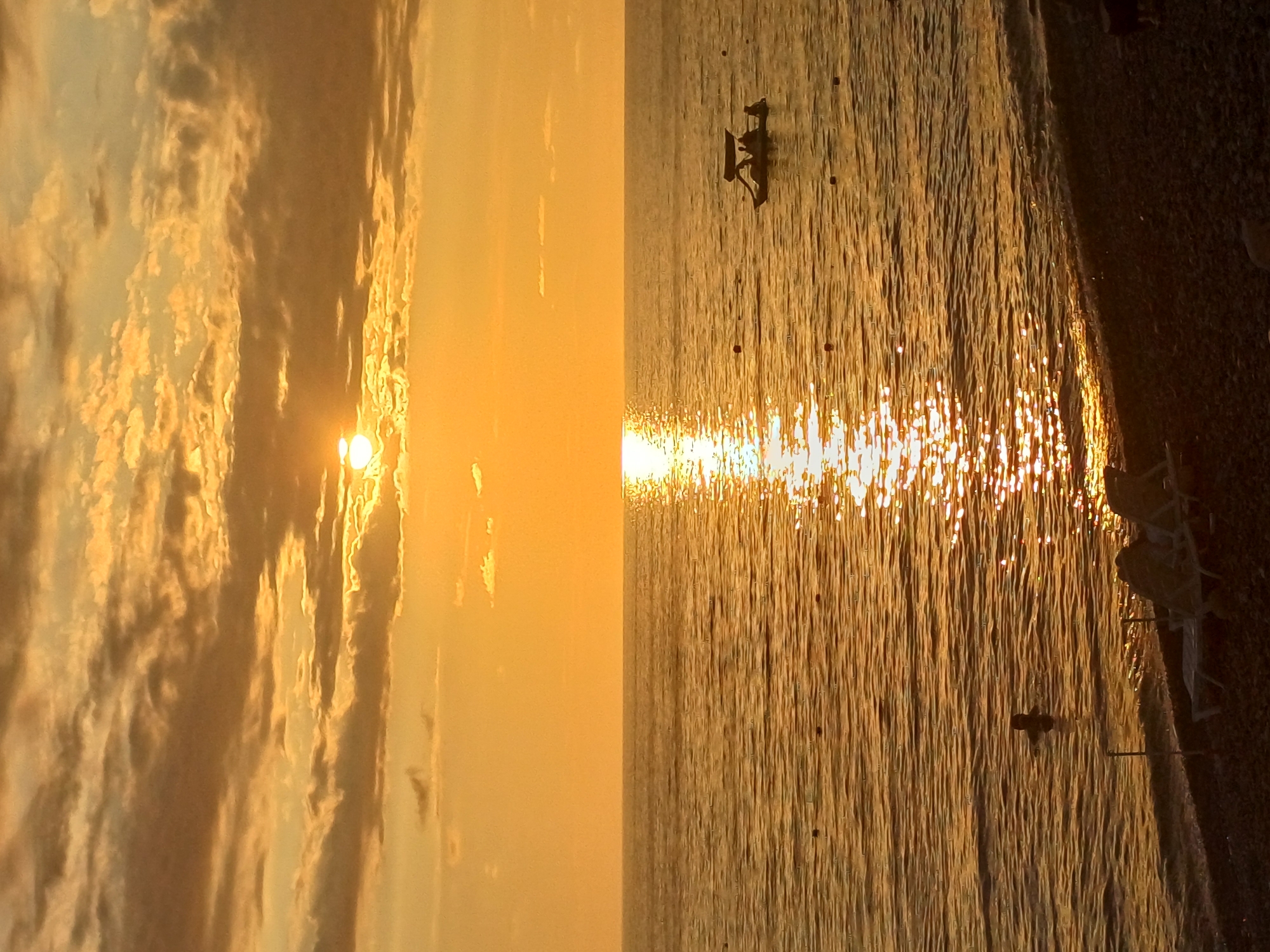 The image depicts a sunset over the Black Sea at a beach in Sochi, Russia. The sky is filled with warm, golden hues and scattered clouds, with the sun partially obscured by the clouds, casting a bright reflection on the water. The sea is calm, with gentle waves lapping at the pebble beach in the foreground.
On the water, a small, dark-colored boat with a canopy is visible, carrying several people. Further out, a lone individual is swimming in the sea. In the distance, there are what appear to be buoys marking a swimming area.
The beach itself is covered in dark pebbles. Several beach chairs are present, suggesting a leisure area. The overall atmosphere is serene and picturesque, characteristic of a sunset on a coastal resort.