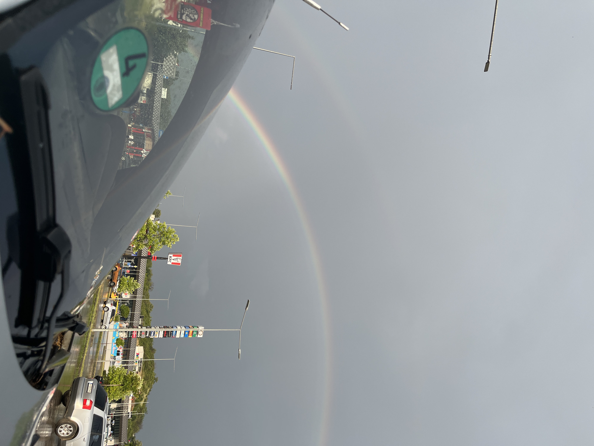 The image captures a cloudy sky over a city street with a prominent double rainbow arcing across the frame. In the foreground, the windshield of a car is visible, along with a green sticker on the windshield displaying the number 4. The background shows a street scene with several vehicles, including a silver station wagon and a yellow hatchback. Buildings line the street, with a KFC restaurant and a tall tower displaying various advertisements visible. Trees with green foliage are scattered throughout the scene. The ground appears wet, suggesting recent rain. The location is identified as Shumen, Bulgaria.