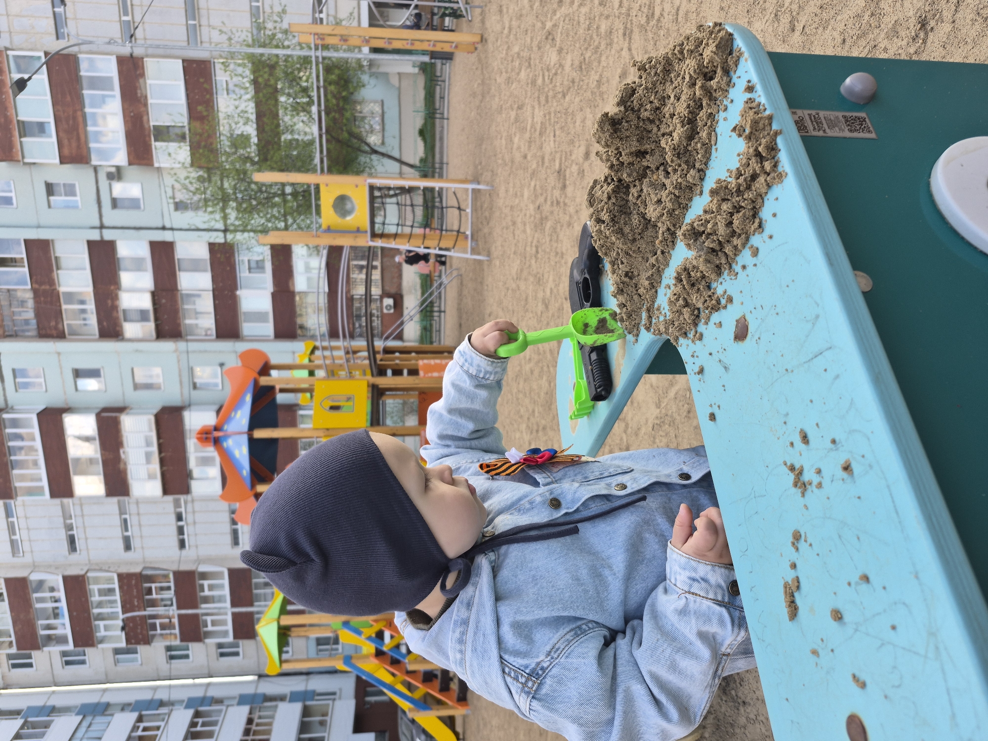 A child in a dark blue cap and light blue denim jacket plays with a green shovel in a sandbox at a playground. The sandbox is light blue with a pile of sand on it. The child has a ribbon pinned to their jacket. In the background, there is a multi-story apartment building. The playground is located in Tobolsk, Russia.