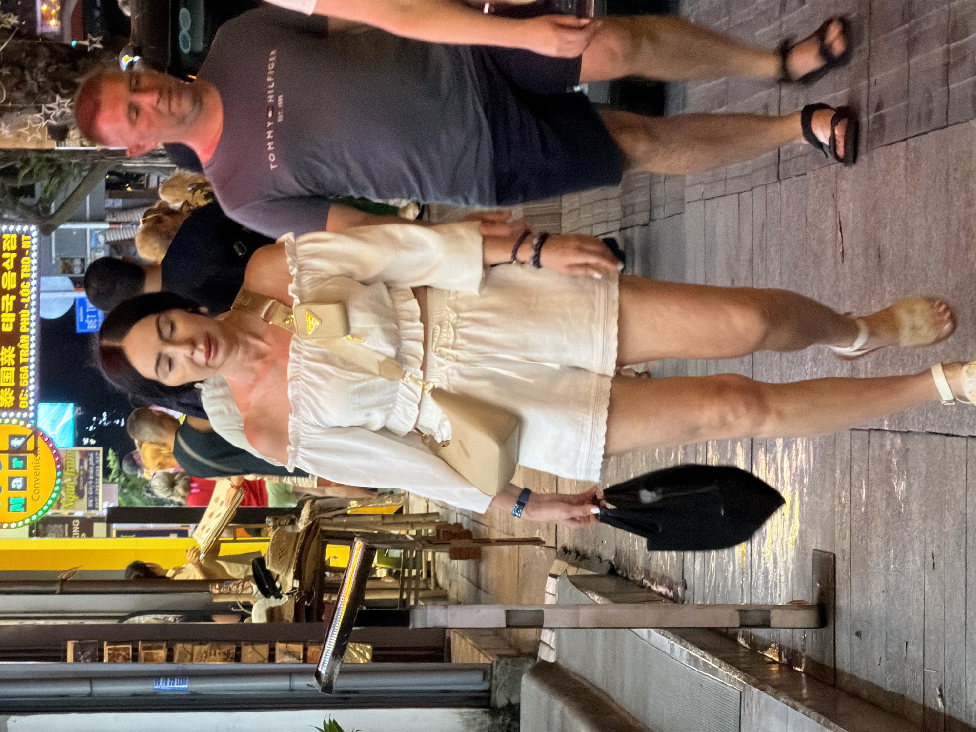 The image shows a street scene in Nha Trang Ward, Vietnam, at night. The foreground features a woman walking towards the viewer, wearing a white off-the-shoulder top and matching shorts. She carries a black bag in her left hand and a small beige purse across her body. To her right, a man walks away from the viewer, wearing a dark t-shirt and shorts. Other people are visible in the background, as are streetlights and signs in Vietnamese. The ground is paved and appears wet, possibly from recent rain.