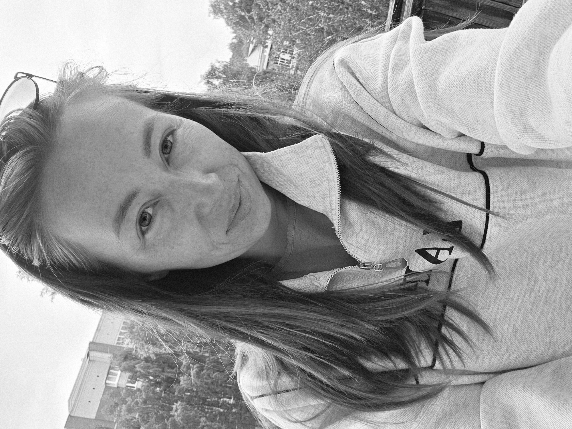 A black and white, eye-level self-portrait depicts an adult female positioned outdoors in Chaikovsky, Russia. She has long, dark hair, worn down with several loose strands, and visible freckles across her nose and cheeks. Dark-rimmed eyeglasses are pushed up onto her head. She is wearing a light-colored (likely grey) quarter-zip sweatshirt with a silver-toned zipper pull. A partial black letter "A" and a horizontal black stripe are visible on the front of her garment. Her expression is a subtle smile, and she looks directly into the camera. The background, viewed from an elevated perspective, features dense deciduous trees. Beyond the trees, a multi-story building with a distinct facade and multiple windows is visible to the far left. A faint wooden railing is present on the right side of the foreground, suggesting a balcony or similar elevated vantage point.