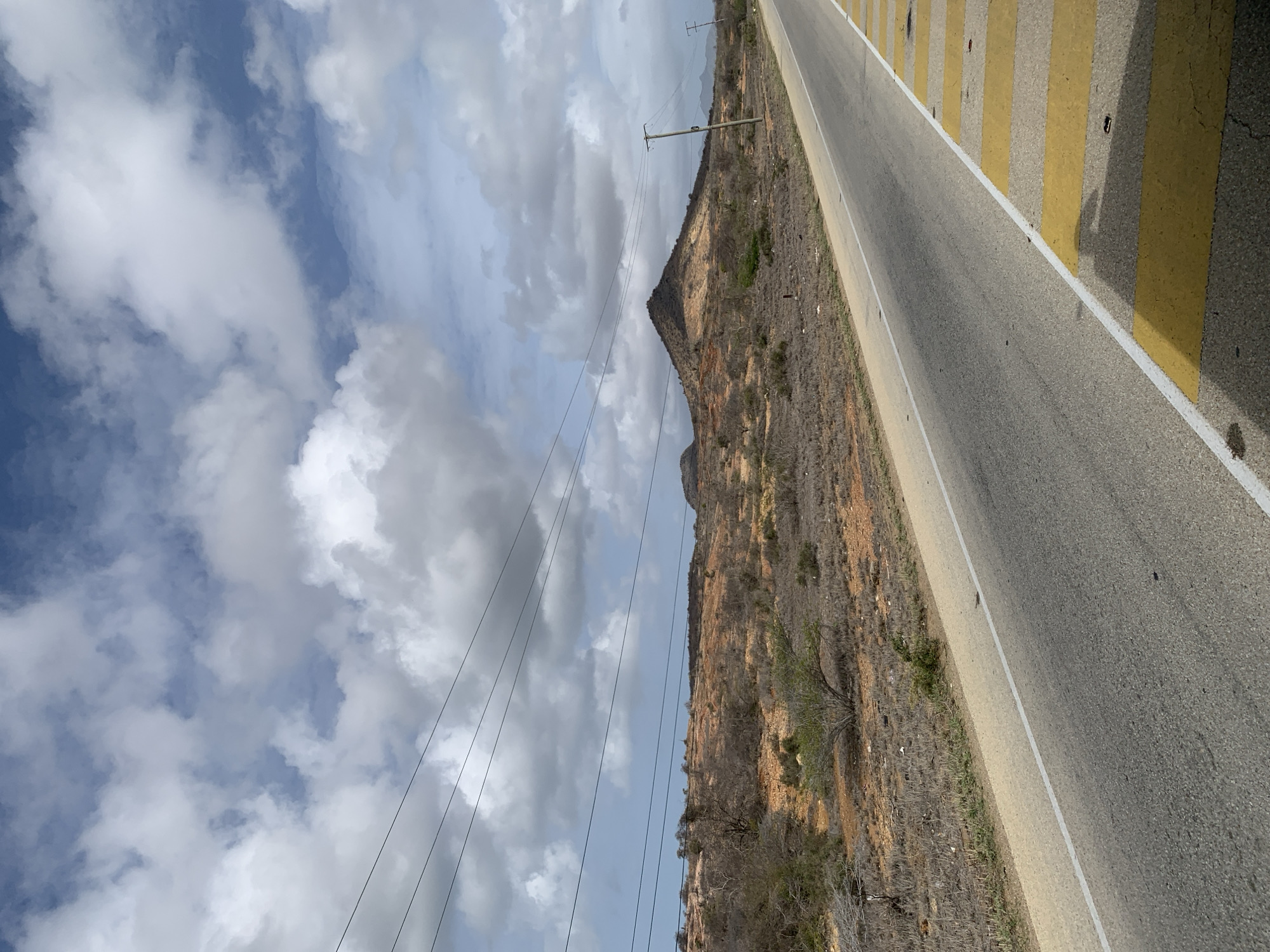 The image depicts a desolate, arid landscape with a paved road running through it. The road has a white line marking the lane and yellow pedestrian crossing markings on the right side. The terrain is dry and sparsely vegetated, with low-lying bushes and scattered trees. In the background, there are rolling hills and a prominent, conical mountain. A few utility poles and overhead wires are visible. The sky is partly cloudy, with a mix of blue sky and white clouds. The scene appears to be in a rural or undeveloped area. The location in Santa María, Venezuela is inferred from the prompt, but not visually evident in the image itself. There are no people or animals visible in the frame, and no specific actions or interactions are depicted.