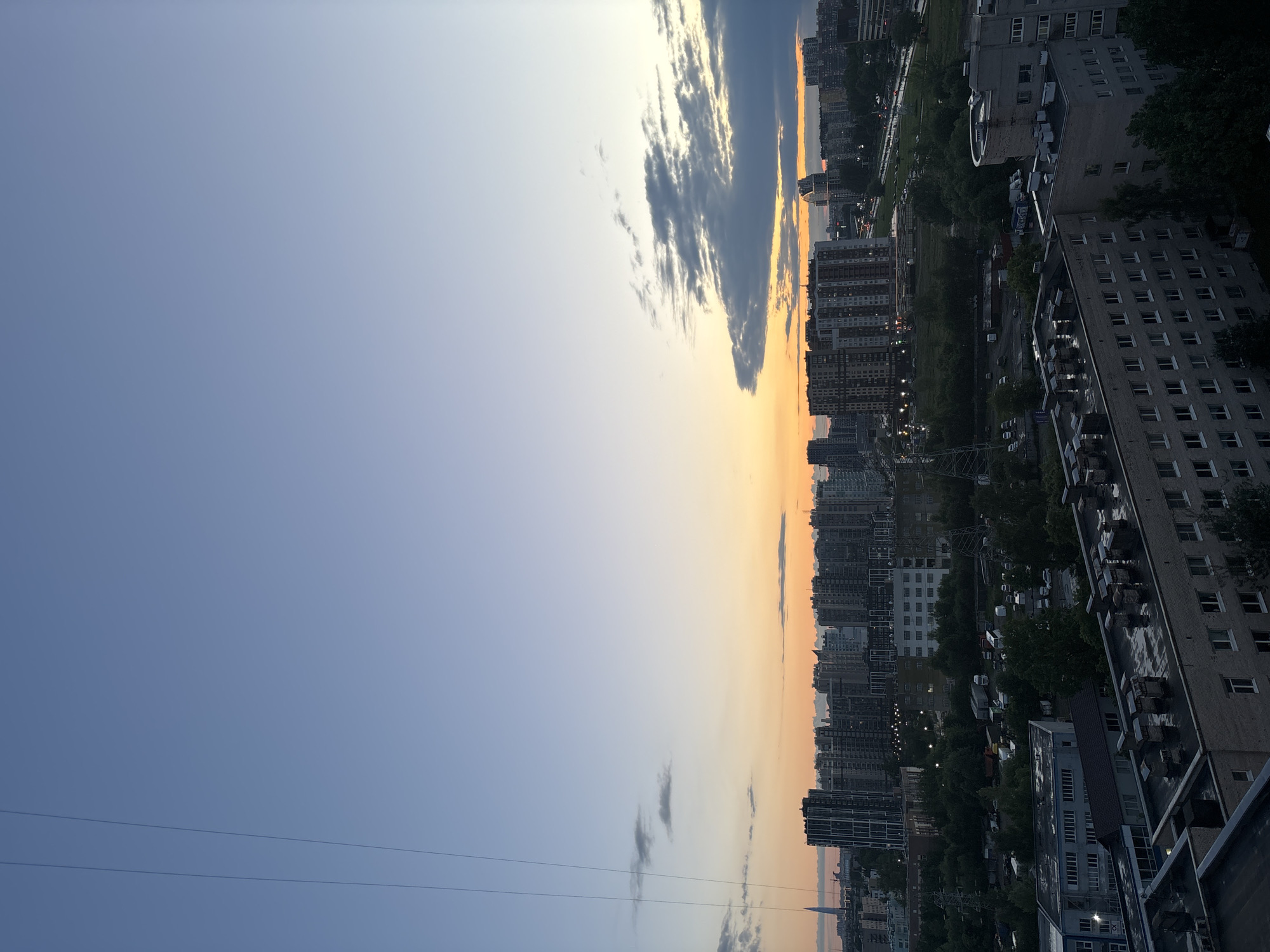 The image is an outdoor, high-angle, full shot of a city skyline at dusk. The sky is a gradient of light blue at the top, transitioning to a soft orange and yellow near the horizon. There are scattered, wispy clouds present. In the foreground, the rooftops of several multi-story apartment buildings are visible. These buildings appear to be constructed of brick and concrete, with visible ventilation systems and satellite dishes on their roofs. Further in the distance, a dense collection of taller, modern apartment buildings forms the city skyline. A few power lines are visible on the left side of the frame. The overall impression is one of a residential urban area during the twilight hours, with no people or active movement depicted. The location is identified as Unknown, Russia.