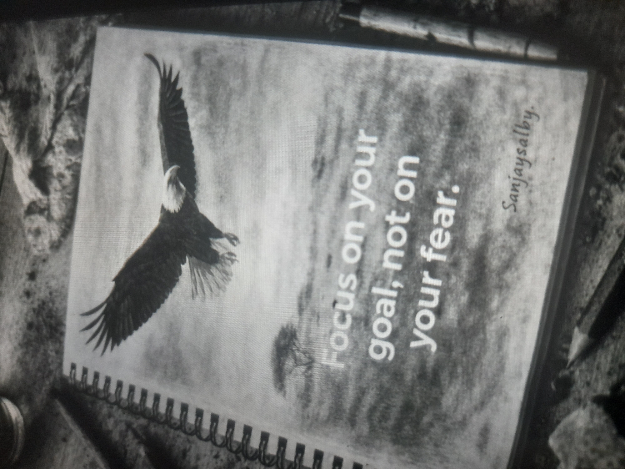 The image is a black and white photo of a notebook with a spiral binding. The notebook lies on a textured surface. The cover of the notebook features an image of a bald eagle in flight and the text "Focus on your goal, not on your fear." The name "Sanjaysalby" is also printed on the cover. Two pencils lie near the notebook. The image does not contain any information that would allow for location within Akwanga, Nigeria.