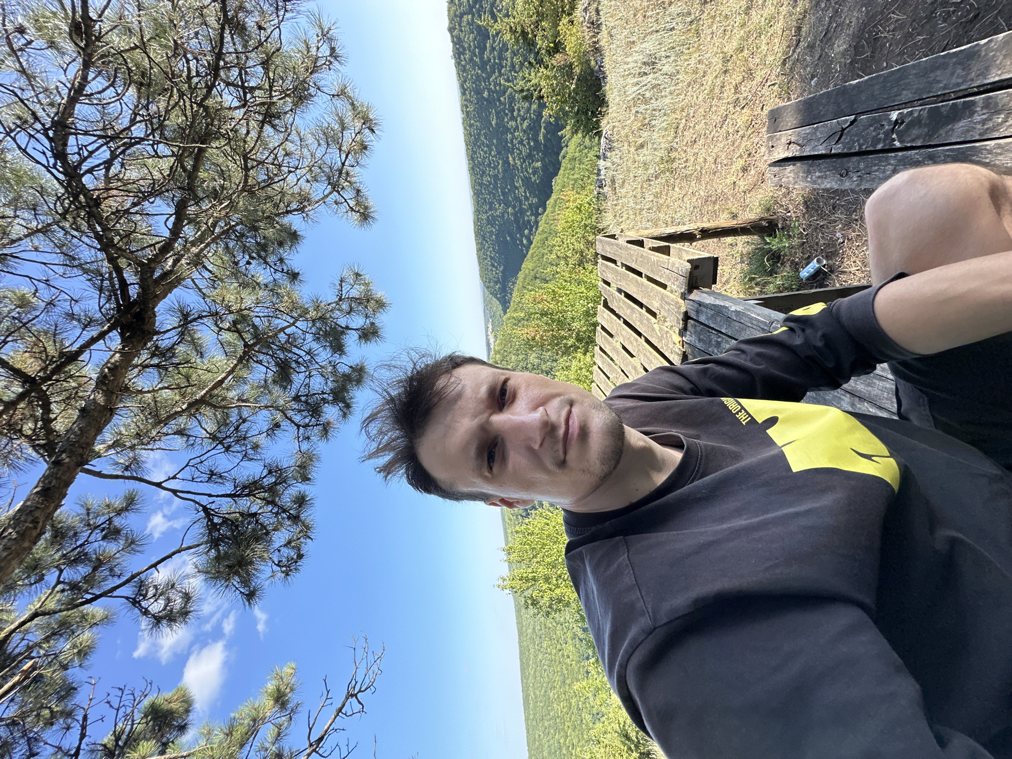A young man with short, dark hair takes a selfie outdoors, sitting on a wooden bench. He is wearing a black long-sleeved shirt with a yellow graphic on the left side of his chest. The background shows a lush green valley with rolling hills covered in trees, under a clear blue sky with scattered clouds. A wooden picnic table is partially visible in the foreground. The setting appears to be a scenic overlook in Pryvorottia Druhe, Ukraine.
