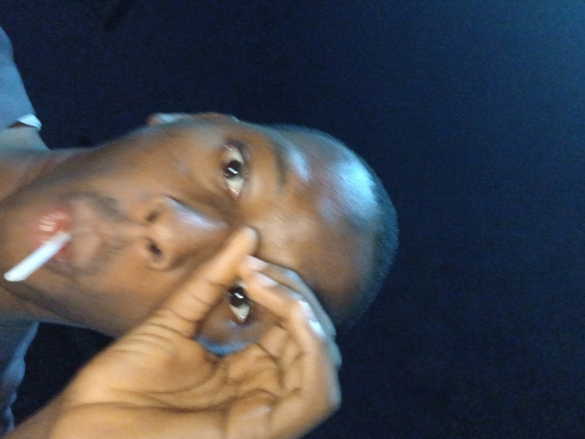A close-up, tilted, indoor shot of a dark-skinned man. The man is holding a white stick in his mouth and has his fingers framing one of his eyes. The background is dark and undefined. There is insufficient information to determine the location within Lagos, Nigeria.