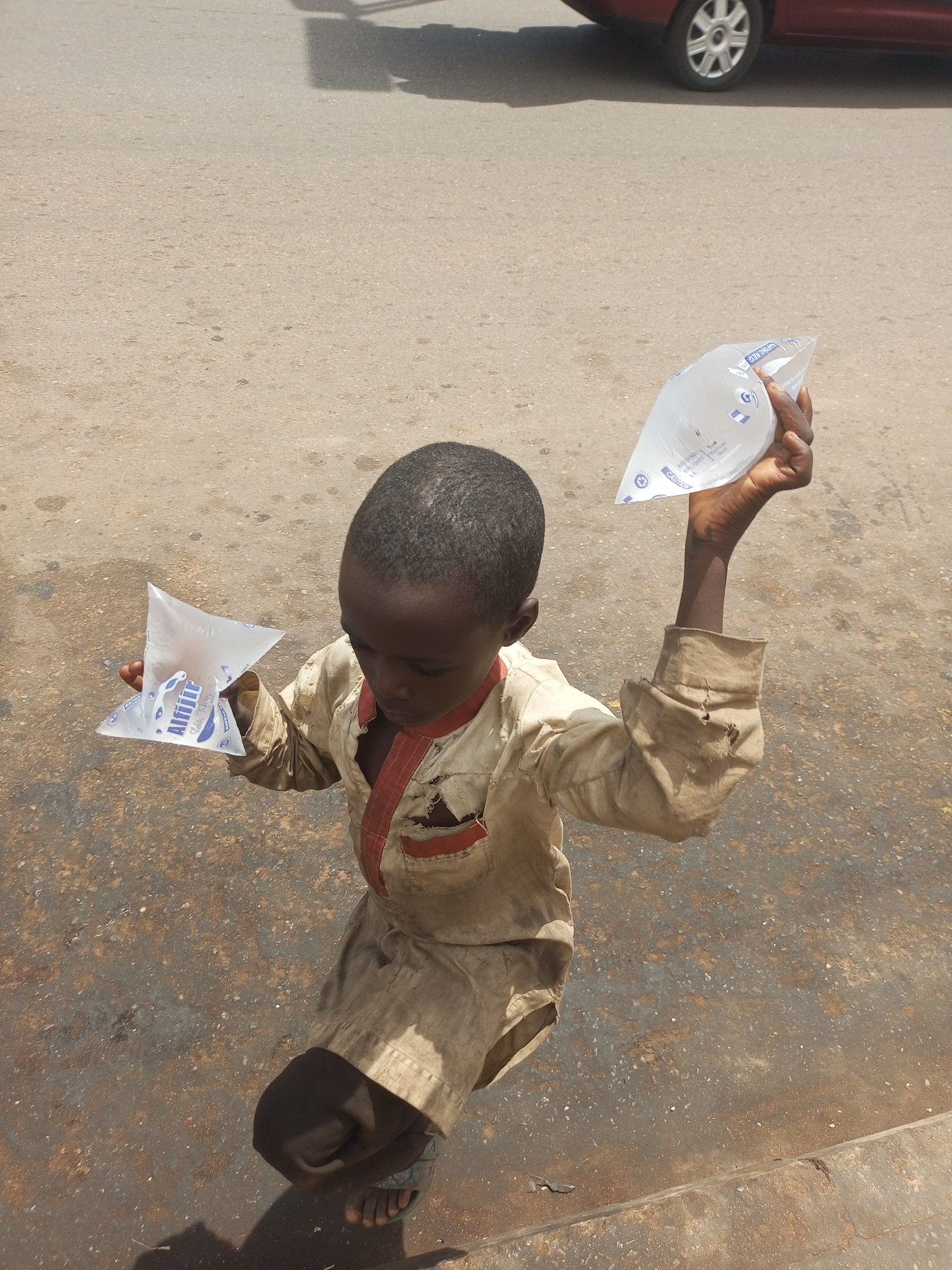 In Gombe, Nigeria, a young boy stands on an asphalt surface, holding two plastic sachets of water. He wears a faded, tan-colored garment with red trim and visible damage on the sleeves and chest pocket. His left hand holds a sachet of water in front of his body, while his right hand, raised and extended, holds another sachet above his head. The ground beneath him is uneven and appears to be a mixture of asphalt and dirt, with scattered debris. In the background, the blurred image of a car, with its wheel visible, suggests an urban setting. The lighting indicates it is daytime, with strong shadows cast on the ground.