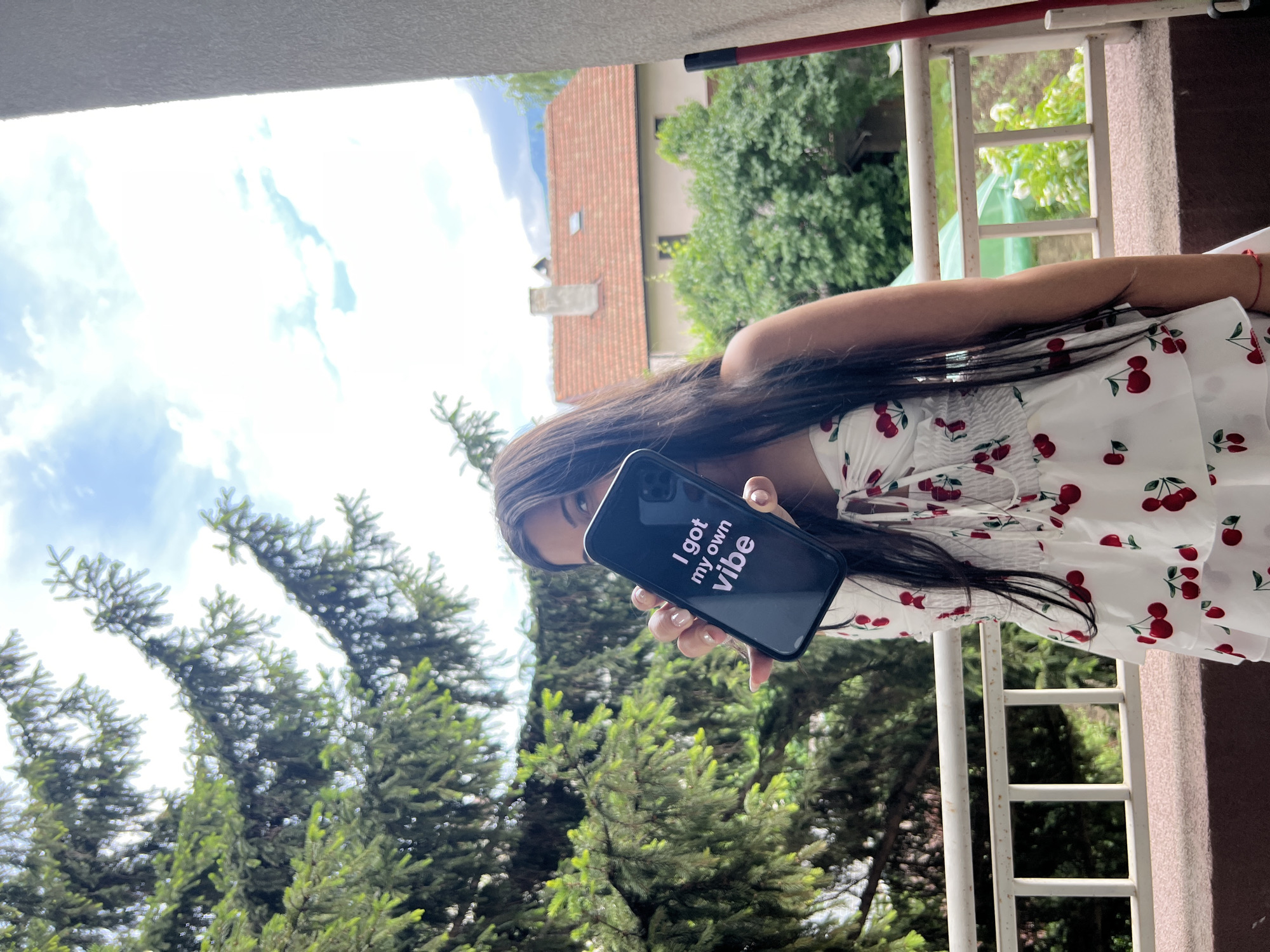 A young woman with long dark hair and a white cherry-print dress stands on a balcony overlooking a residential area with houses and trees. She is holding a smartphone in her right hand, displaying the text "I got my own vibe." The background includes a large evergreen tree, a building with a red tiled roof, and a cloudy sky. The location is believed to be Sofia, Bulgaria, given the architectural style of the houses and the surrounding vegetation.