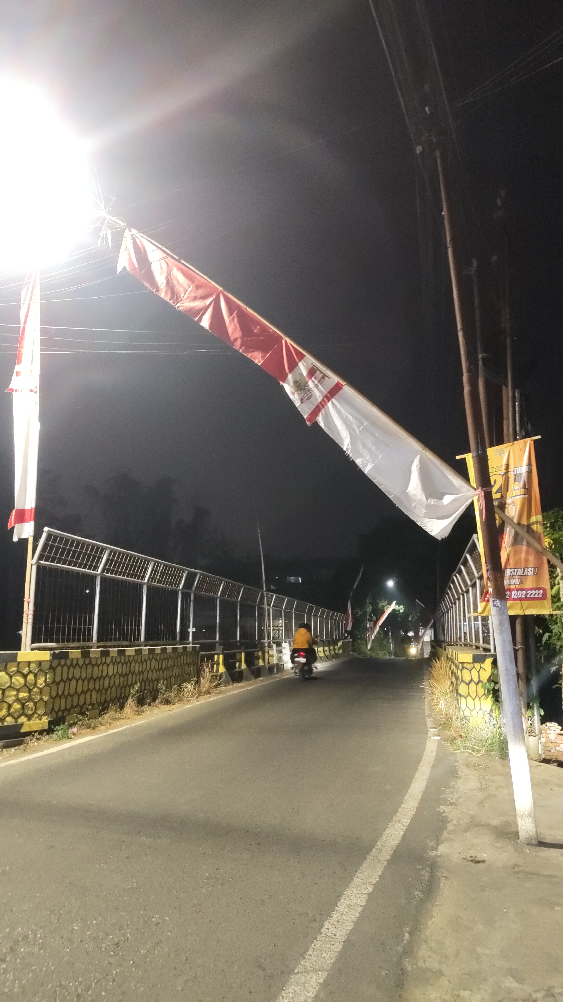 The image shows a road with a bridge, decorated with Indonesian flags, at night. A person on a motorcycle is riding on the road. The bridge has a metal railing and a yellow and black patterned border. There are streetlights and power lines overhead. A banner advertising installation services is visible on a pole. The overall setting appears to be a semi-urban or rural area in Malang, Indonesia.