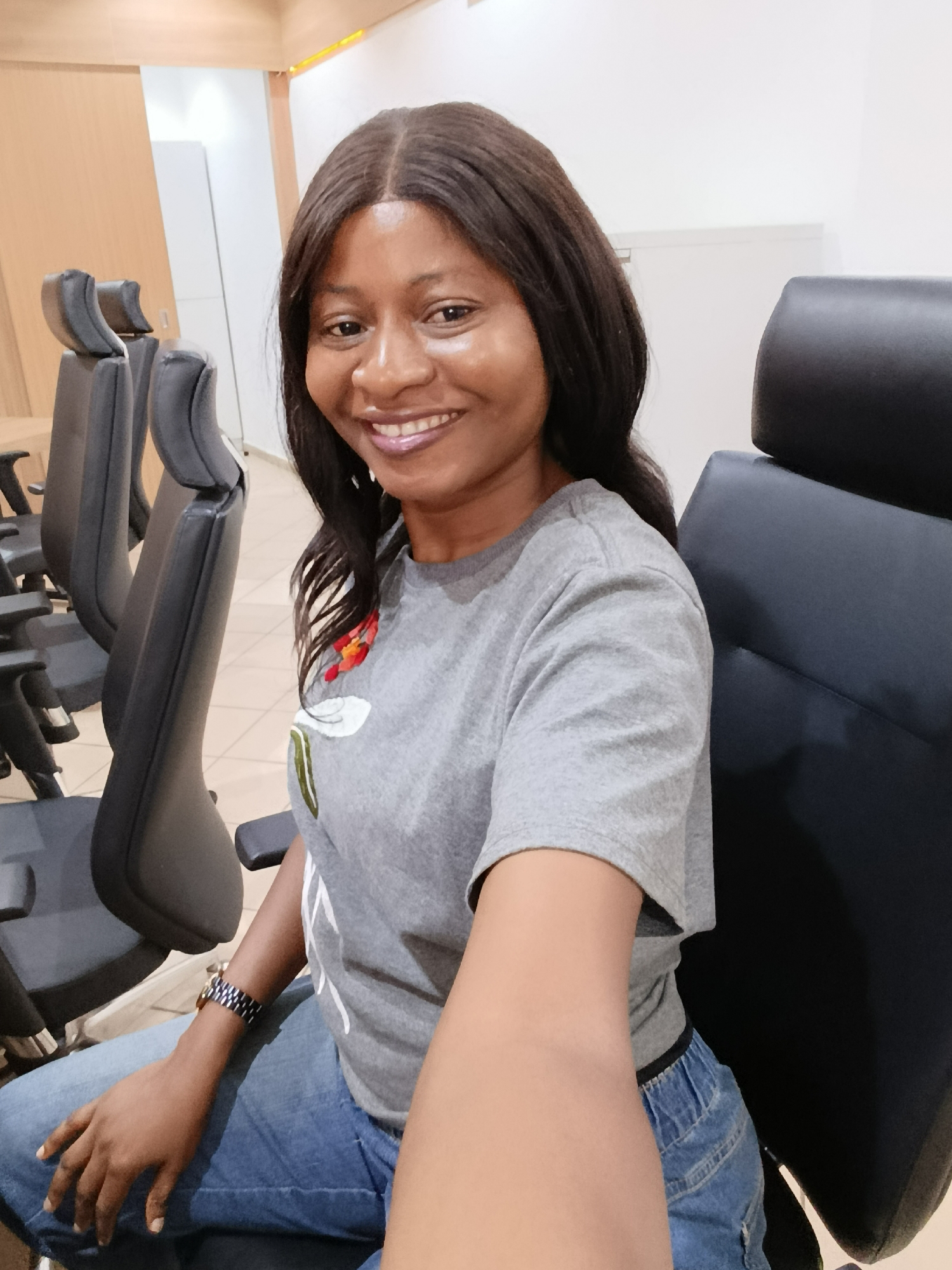 A medium shot depicts a dark-skinned woman with shoulder-length dark hair sitting in a black office chair. She is smiling directly at the camera. She is wearing a gray t-shirt with an embroidered floral design on the upper left side and blue jeans. A wristwatch is visible on her left wrist. She appears to be holding a phone to take a selfie. Behind her are several other black office chairs in a row. The location appears to be an office setting with light-colored walls and a door partially visible in the background. The location is Immigration Quarters, Nigeria.