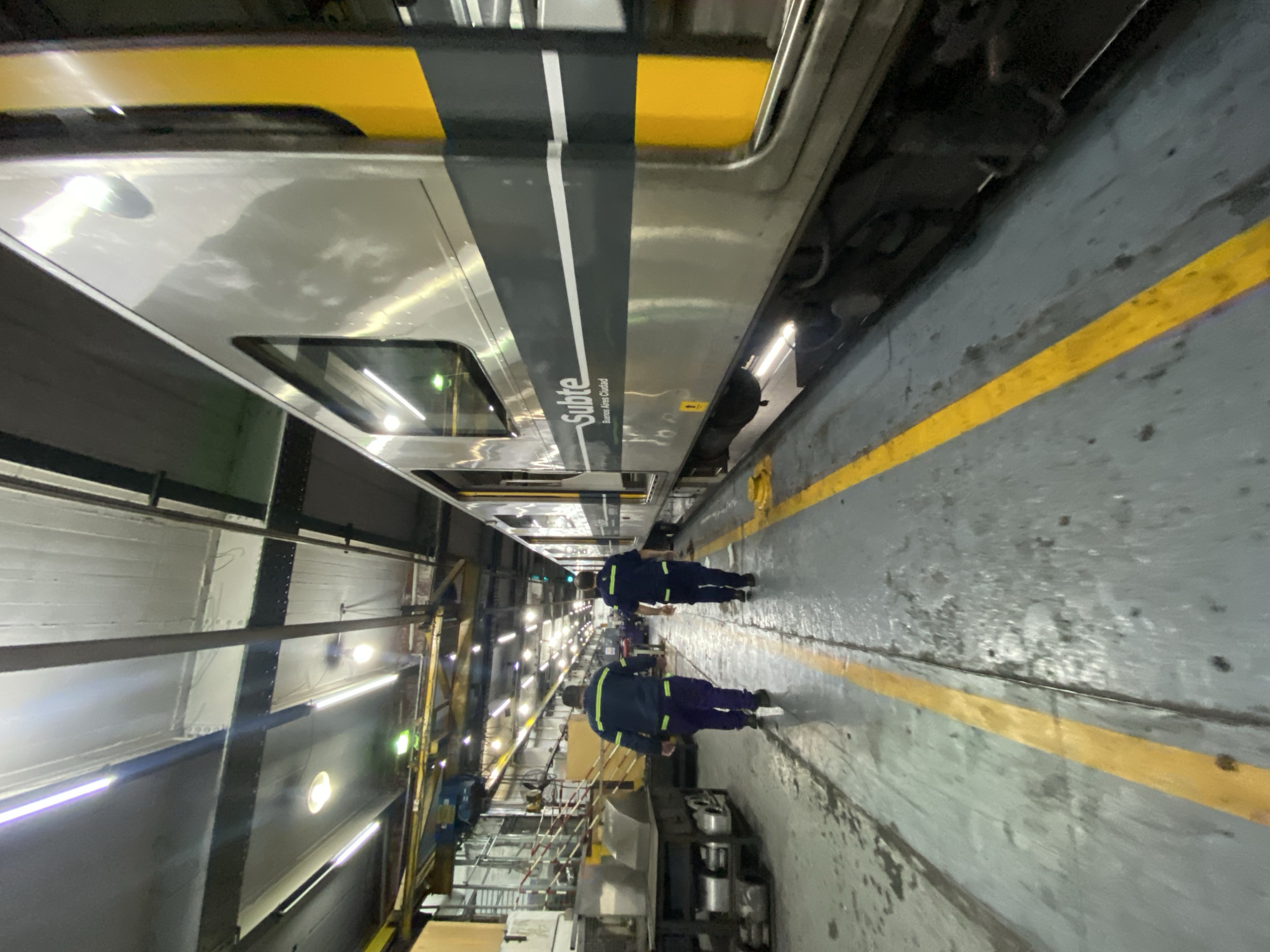 The image shows the interior of a train repair facility in Buenos Aires, Argentina. A silver train car, marked "Subte Buenos Aires Ciudad," is overhead. Two workers in navy blue uniforms with reflective stripes are walking in the foreground. The floor is gray concrete with a yellow safety stripe. Equipment and industrial storage racks are visible along the walls of the facility. Fluorescent lights illuminate the area.