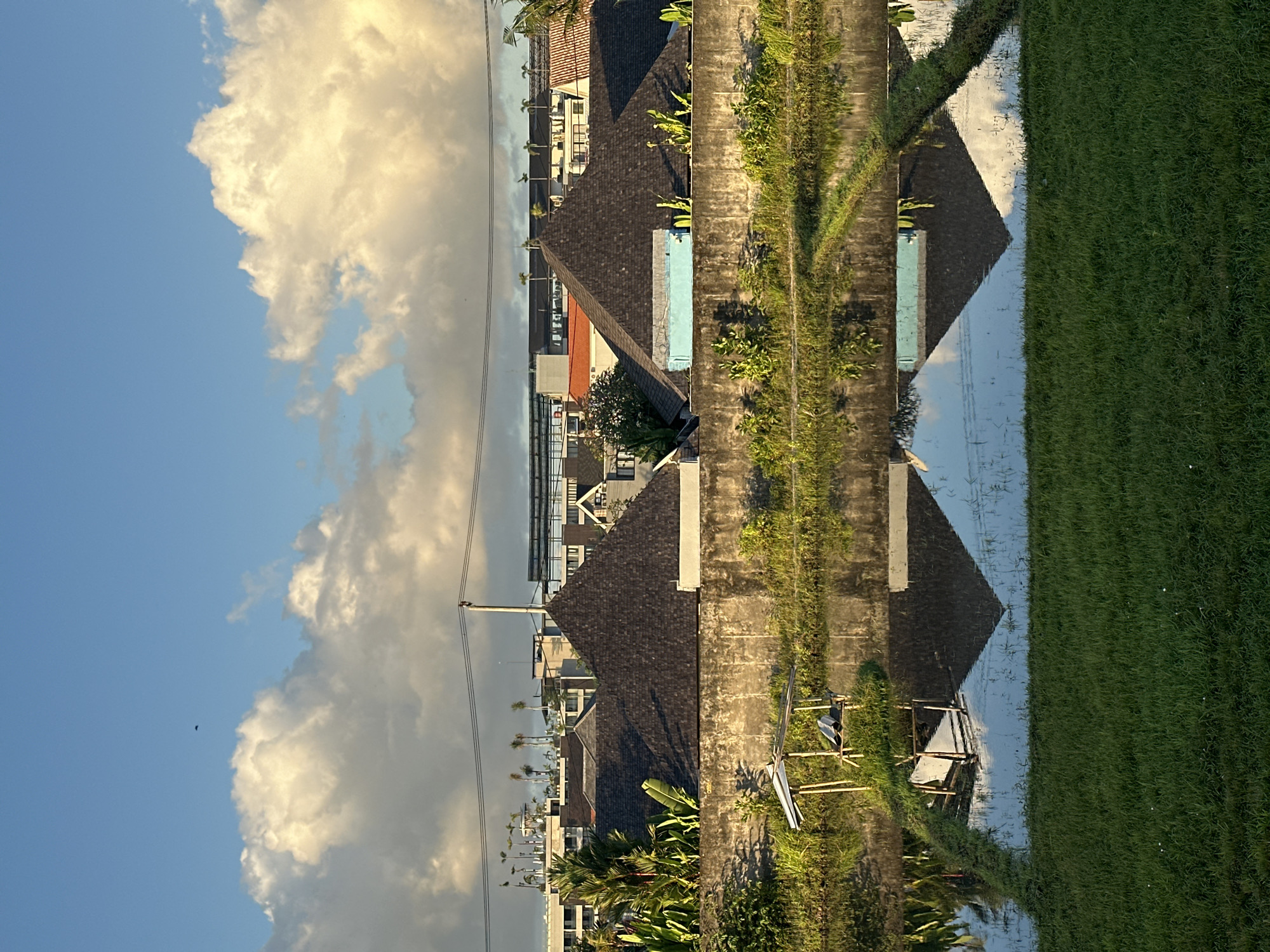 The image shows a scene in North Kuta, Indonesia. Buildings with dark, gabled roofs line one side of a concrete path. Lush greenery and rice fields are visible on the opposite side of the path. The buildings are reflected in the water-filled rice paddies. A wooden structure with metal components is located near the path. The sky is partly cloudy. Power lines are present.