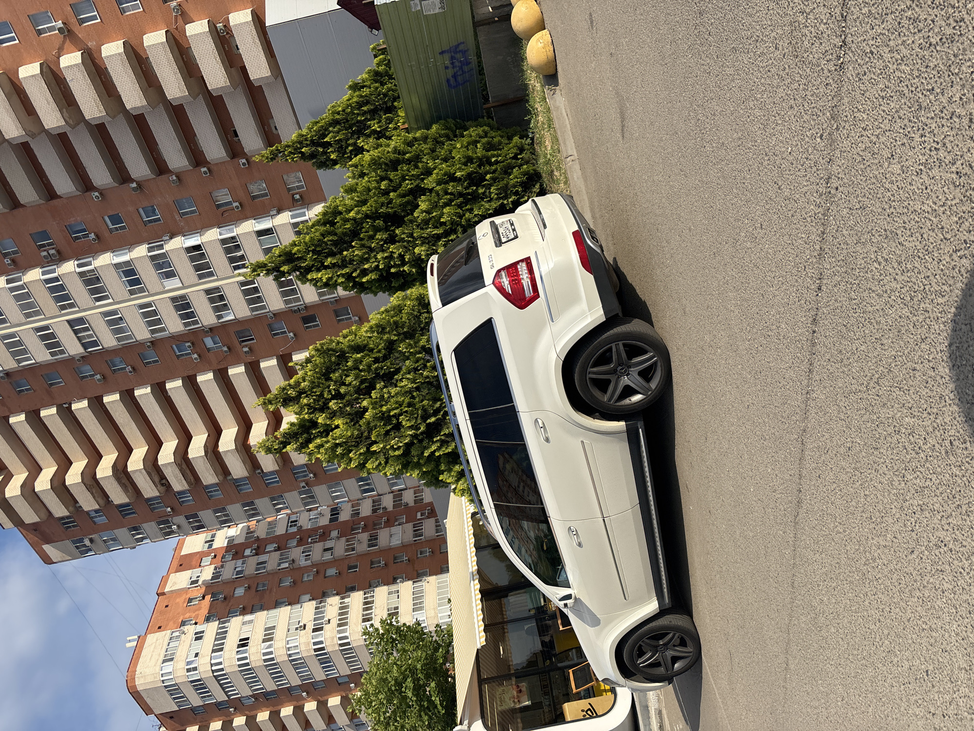 A white Mercedes-Benz SUV is parked on an asphalt street in front of a building. The building appears to be an apartment complex with many windows and balconies. There are also some trees and a green fence visible. The sky is blue, suggesting it is daytime and likely sunny. The car is the primary subject, positioned in the foreground and occupying a significant portion of the image. The angle of the photograph is from a slight distance, looking towards the rear of the vehicle. The image captures a common urban scene. The license plate of the car is partially visible, displaying Cyrillic characters and numbers, consistent with Russian license plates. There is also some graffiti on the green fence behind the car. The overall impression is a casual snapshot of a car parked in a residential area.