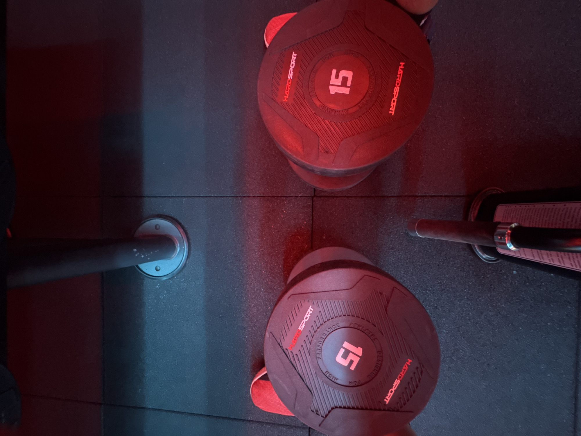 In the city of Almaty, Kazakhstan, a close-up, high-angle shot shows two red and black dumbbells, each marked with the number "15" in a circle. The dumbbells are positioned on a dark, textured floor, likely in a gym setting, as indicated by the presence of a metal support pole with a circular base. The lighting is predominantly red, with some blue light visible in the background, suggesting artificial illumination. The dumbbells are the primary focus, with their textured surfaces and prominent weight markings clearly visible.