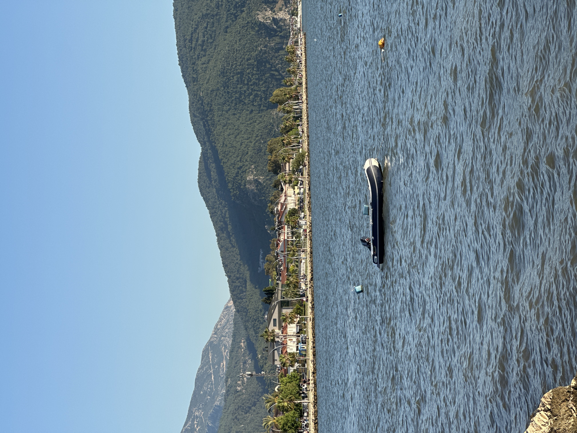 The image shows a coastal scene in Fethiye, Turkey. A dark-colored inflatable boat with one person aboard is in the water near the shoreline. Along the coast, there's a line of buildings, trees (including palm trees), and streetlights suggesting a developed promenade or road. A steep, forested mountain rises behind the buildings, and the sky is clear and blue. Several buoys are floating in the water near the boat. The water surface has visible ripples, indicating a light breeze or current. The view is primarily focused on the boat and the shoreline, with the mountain serving as a backdrop. The scene suggests a sunny day with calm conditions for boating.