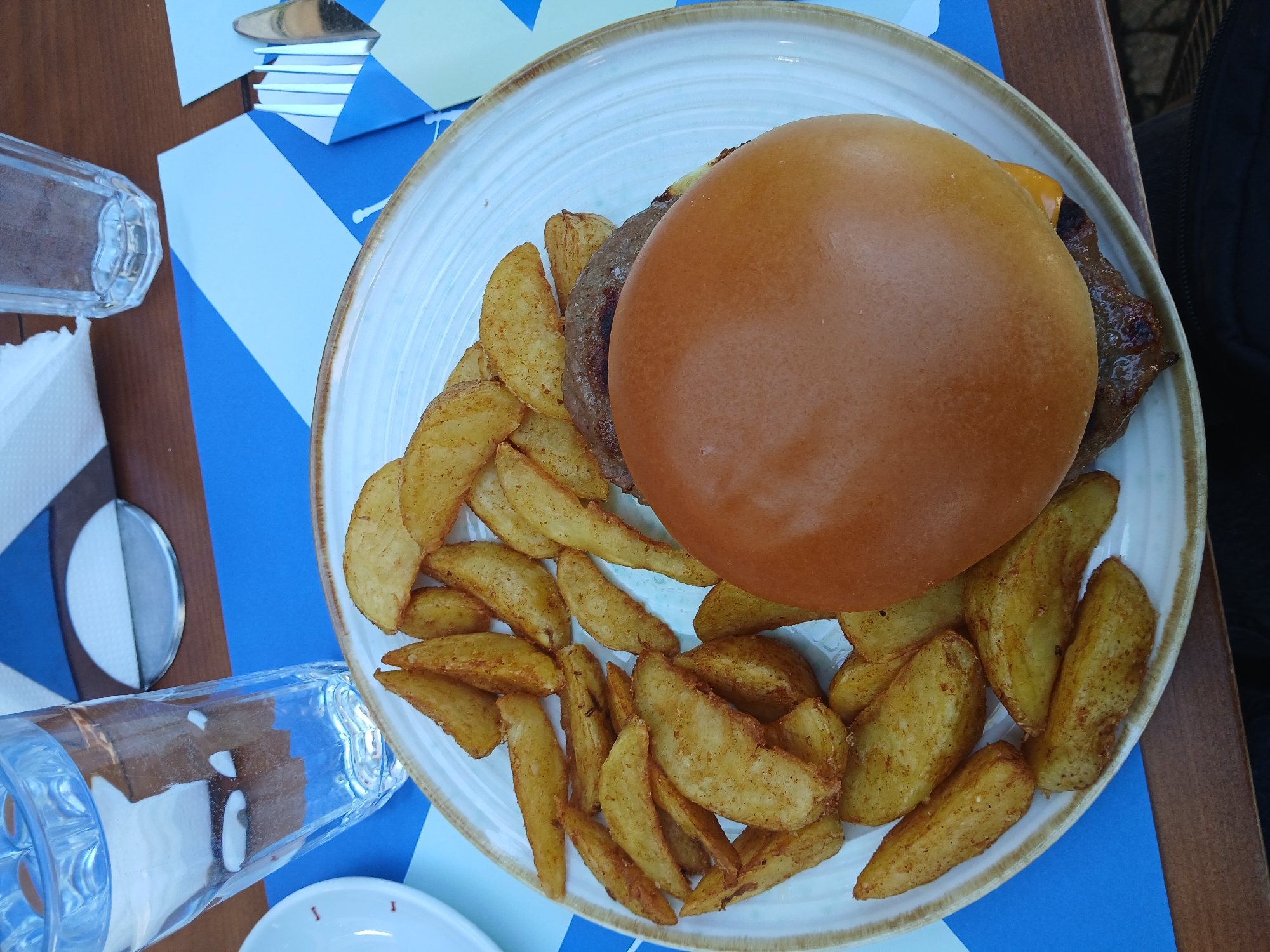 A close-up, overhead shot shows a plate of food on a table. The plate contains a hamburger with a golden-brown bun and a side of potato wedges. The food is presented on a white plate with a textured rim. To the left of the plate is a glass of water, and to the right, a fork is visible tucked into a blue napkin. The table surface is covered with a blue and white patterned placemat. The background suggests an outdoor or casual indoor dining setting, possibly in Cetinje, Montenegro, indicated by the blue and white geometric paper placemats, common in European cafes and restaurants. The lighting is natural, highlighting the textures of the food and the glass.