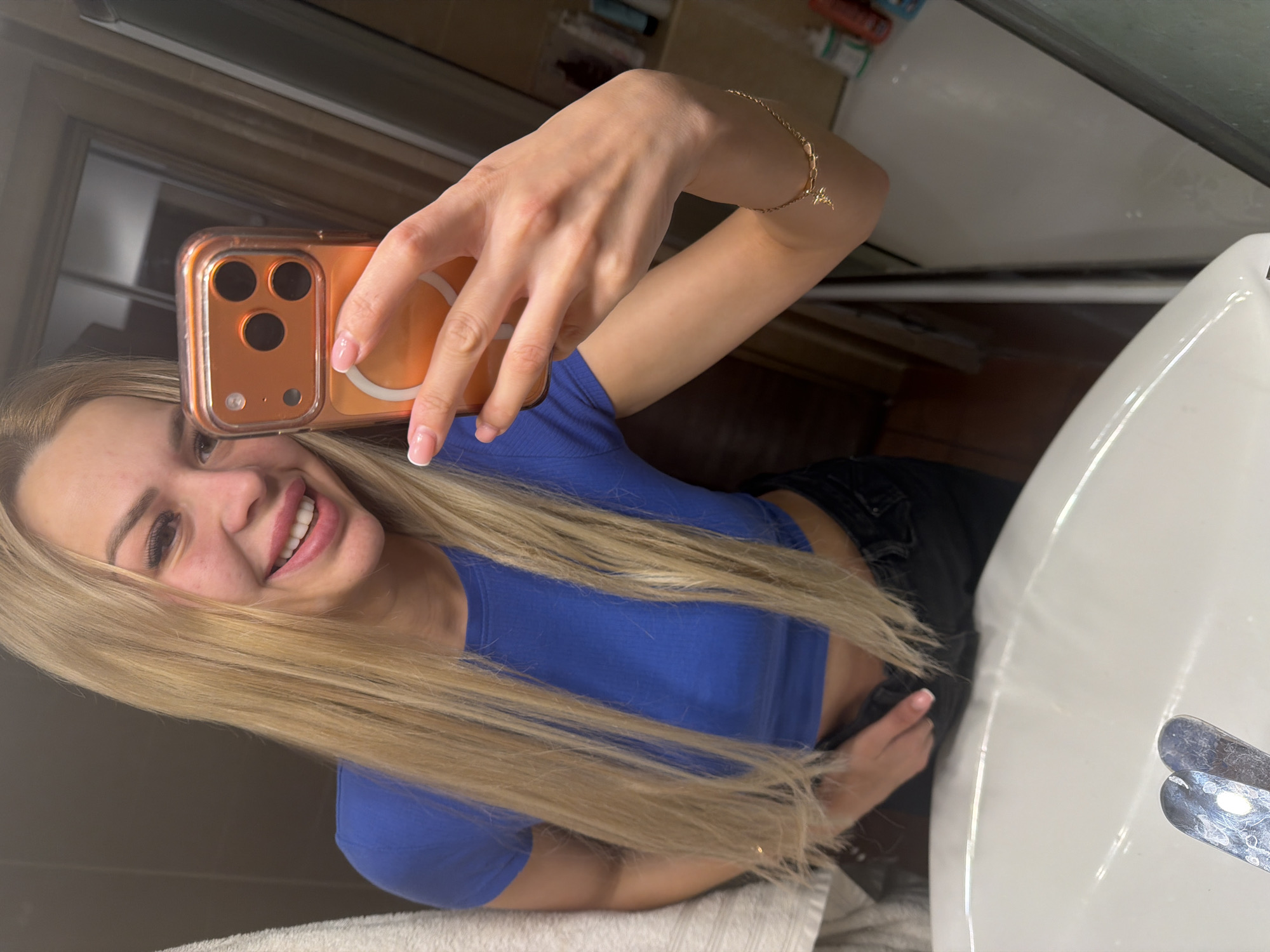 A woman with long blonde hair is taking a selfie in a bathroom mirror. She is wearing a blue crop top and black pants. She has on makeup and her nails are painted pink. A gold bracelet is on her right wrist. The background of the image shows a bathroom sink, a towel on the left, and a door and shelves in the background. The mirror reflects the woman, the bathroom, and a glimpse of a hallway. It is not possible to determine the location in the city of Kyiv, Ukraine from the image alone.