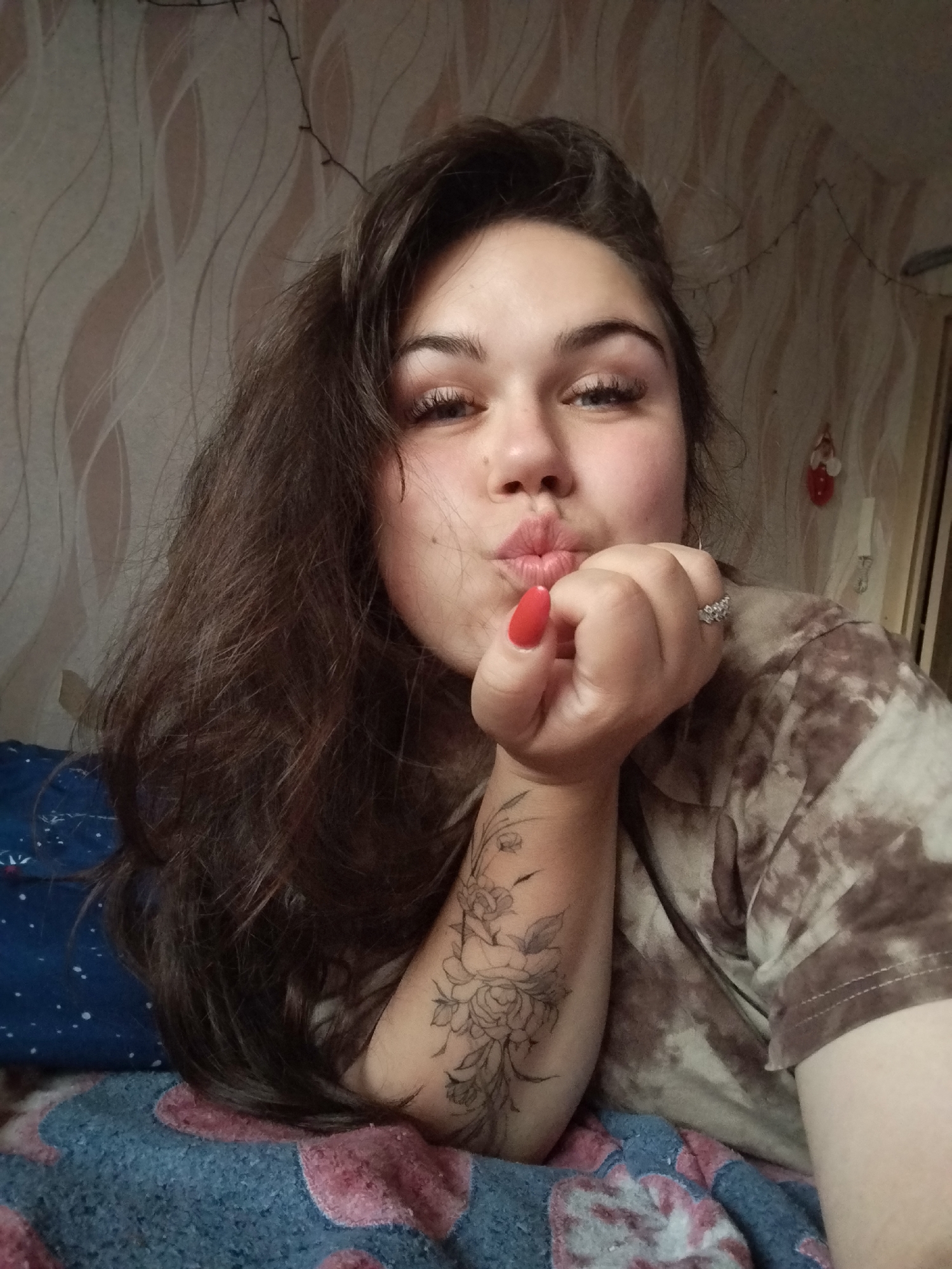 A young woman is captured in a close-up, eye-level selfie, striking a playful pose indoors. She has long, dark brown hair and light-colored eyes, with prominent eyelashes and well-defined eyebrows. Her lips are puckered in a "kissing" gesture as she rests her chin on her right hand. Her fingernails are painted a vibrant red, and a silver-toned ring with multiple small stones adorns her right ring finger. A detailed black and white floral tattoo, featuring roses and other blossoms, is visible on her right forearm.
She is wearing a casual t-shirt with a brown and beige tie-dye pattern. The woman appears to be reclining on a bed or sofa, supported by a blue pillow with white speckles, and a blue and pink patterned blanket is visible beneath her. The background features a wall covered in light-colored wallpaper with a subtle, wavy textured design. A string of delicate lights is draped across the top of the wall, and a small, red and white ornamental hanging can be seen below it. The setting conveys a relaxed, personal atmosphere, likely within a bedroom. No text is visible in the image. The photo was taken in Chaikovsky, Russia.