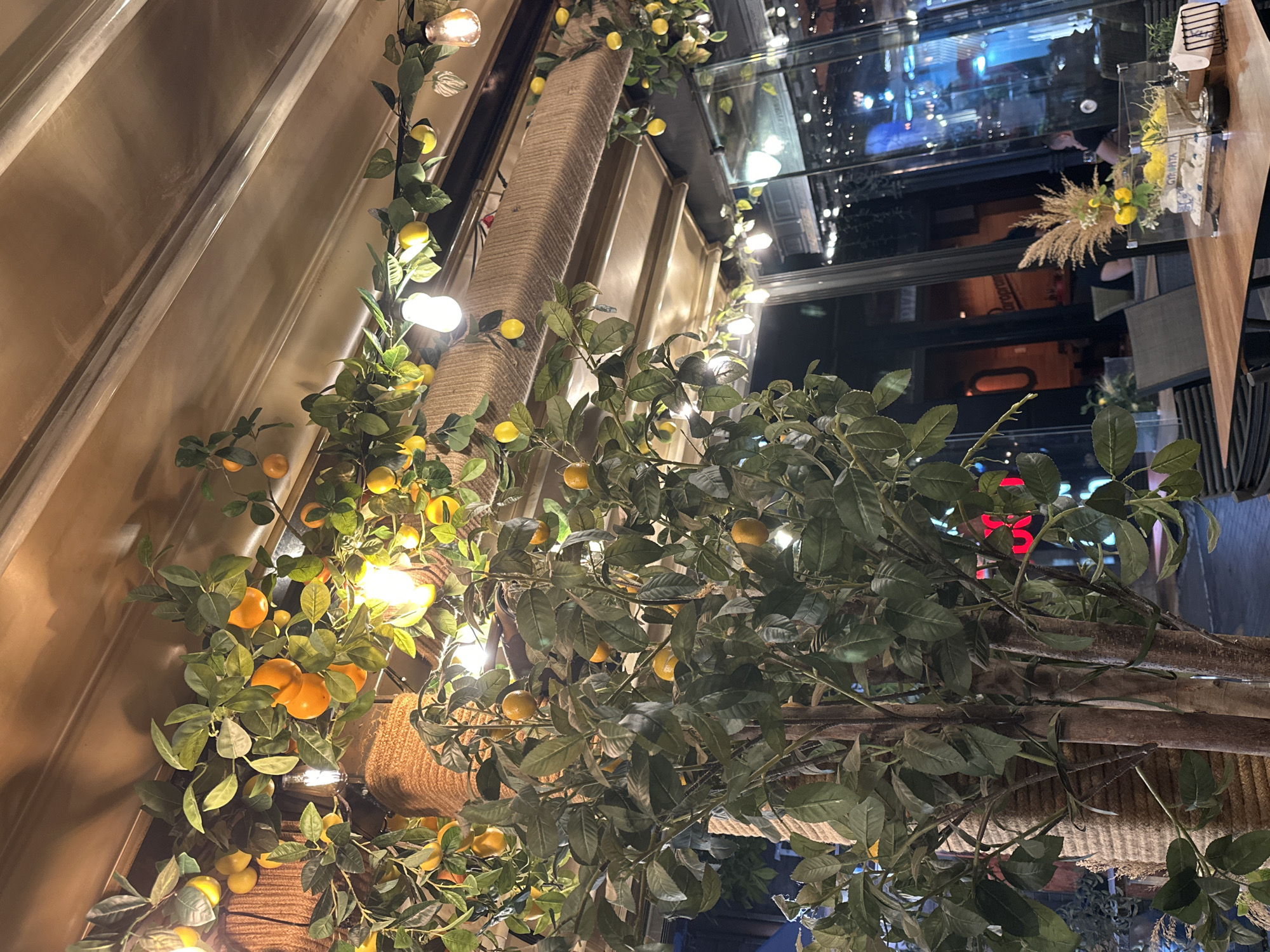 An indoor scene in what appears to be a restaurant or cafe is captured in the image. Artificial fruit-bearing branches, with green leaves and orange and yellow fruits, are draped along the ceiling and a central pillar. Small, decorative lightbulbs are interspersed with the foliage. In the background, through large glass windows, a city scene is visible with blurred lights and reflections. A table with a decorative centerpiece featuring yellow flowers and rocks is in the foreground. Two individuals are partially visible through the glass, seated at tables in the background. The overall ambiance suggests a warmly lit, possibly evening setting.
