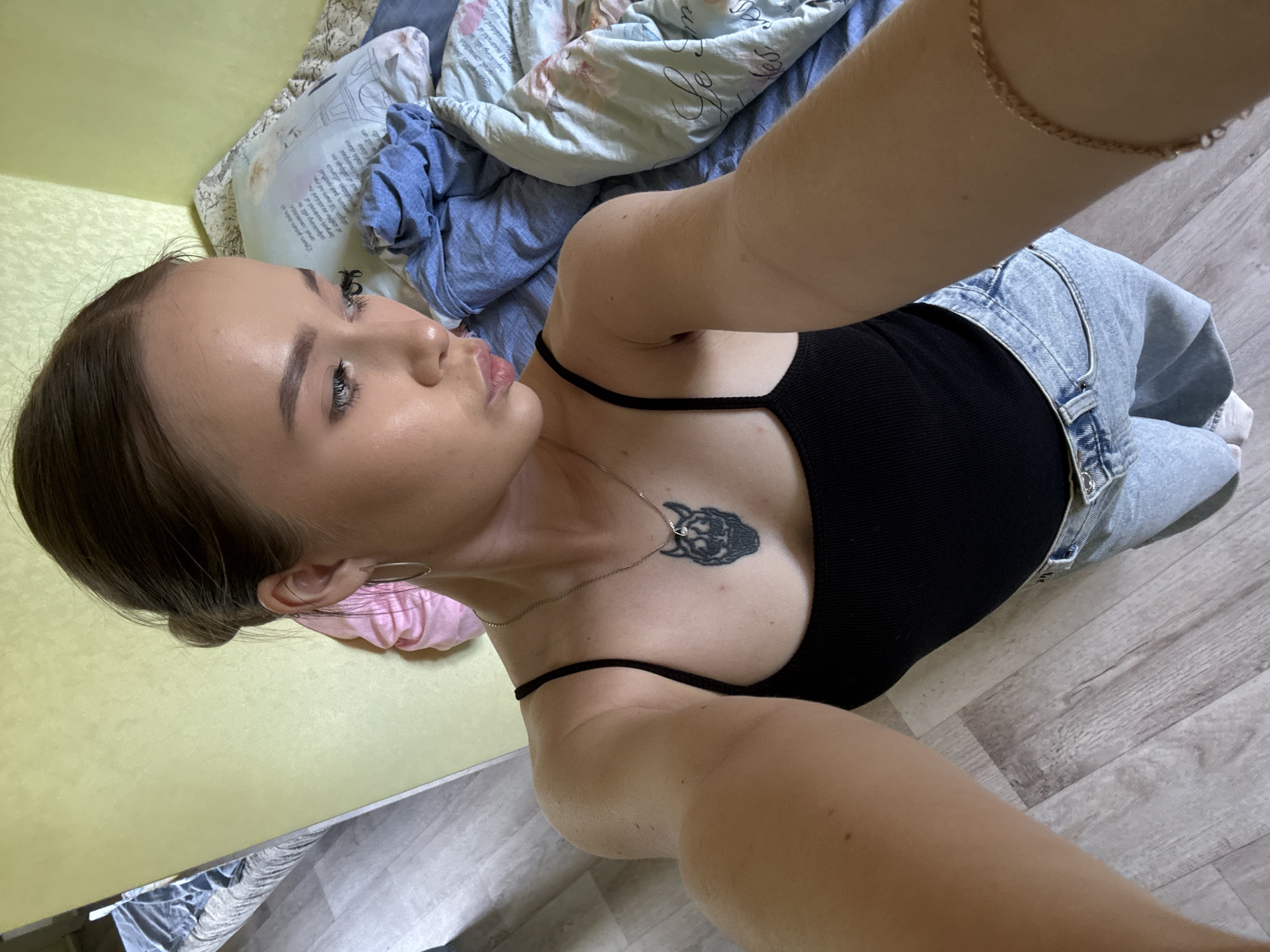 A woman with fair skin and brown hair pulled back into a low bun is lying on her back on a bed or floor in an indoor setting. She is wearing a black tank top and light blue jeans. A small tattoo is visible on her chest, near her left clavicle. The woman's face is turned to her left. She is wearing earrings and a necklace. The environment includes bedding with a mix of blue and floral patterns. The floor is covered with light-colored wooden planks. The wall behind her is a light yellow. The location is Unknown, Russia.