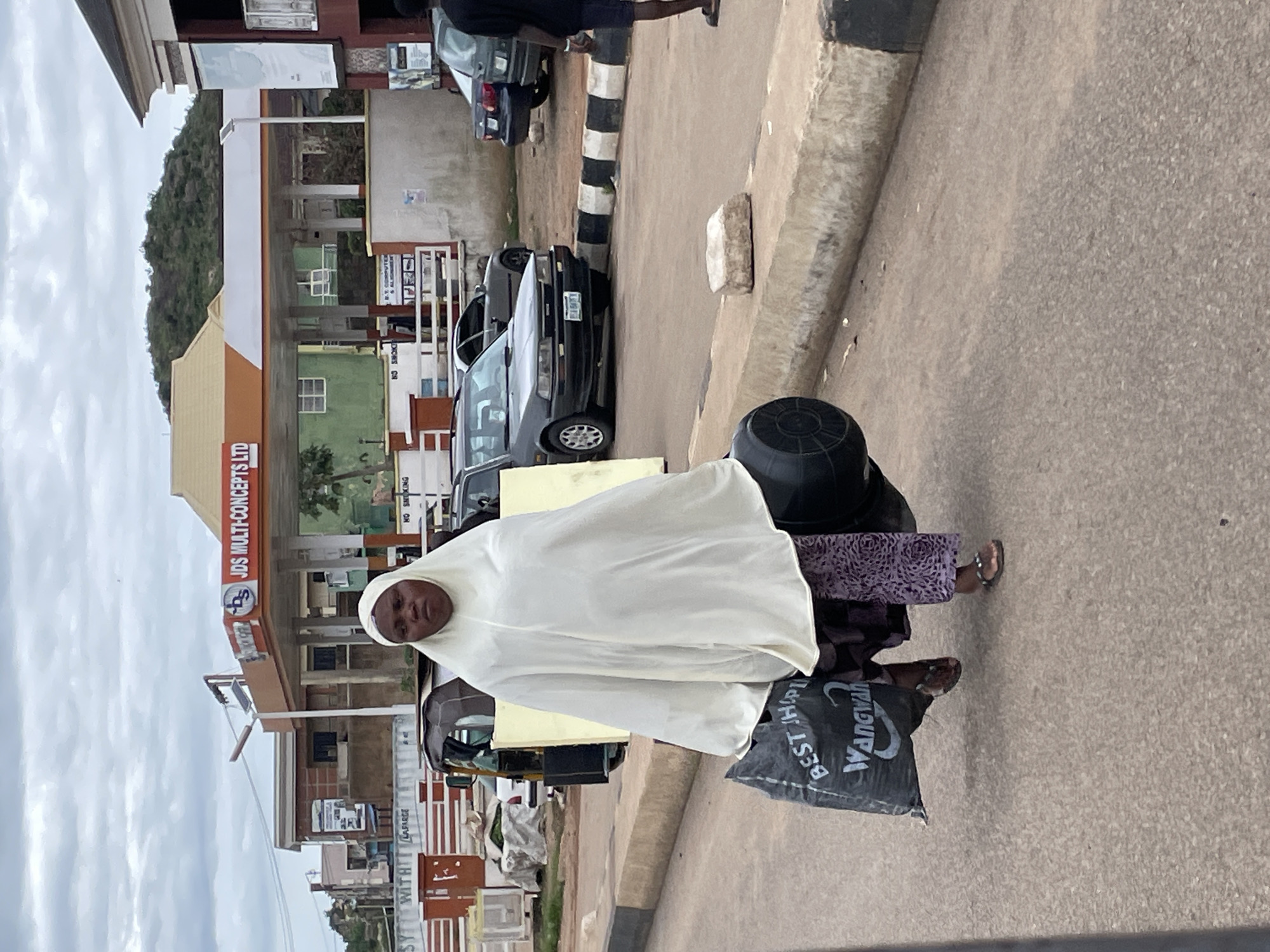 This image captures a scene on a street in Gura Top, Nigeria, likely during daylight hours given the bright sky and shadows. The primary subject is a person wearing a white hijab and a patterned skirt, standing near the roadside. They are carrying a large black bag with text on it, and a dark bucket is visible behind them. The person's expression appears neutral.
The setting is urban, with buildings and vehicles present. A prominent building with signage is to the left, displaying "JDS MULTI-CONCEPTS LTD" and a logo. Several cars are parked or moving on the street, including a dark sedan in the foreground and other vehicles further back. A striped pole, likely a traffic marker, is also visible. The ground is paved with concrete.
The overall atmosphere seems to be that of an ordinary day in a busy area, with the individual seemingly engaged in daily activities, possibly transporting goods. The sky is overcast, suggesting a cloudy day. The visible text on the black bag includes "BEST JHL" and "WANGWANA", though the full context is unclear.