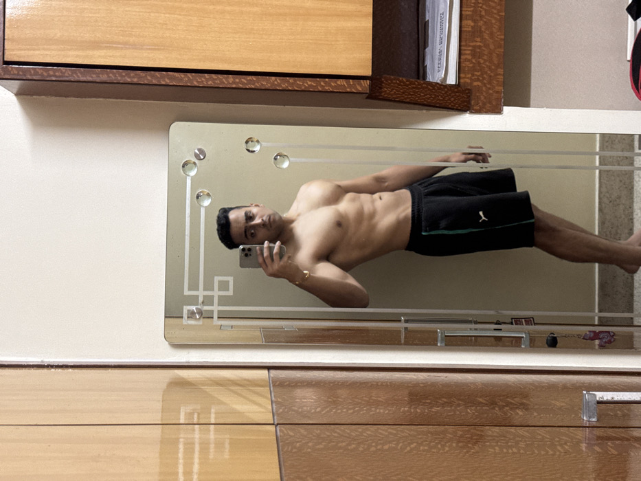 A shirtless man is reflected in a mirror, holding a cell phone, presumably taking a selfie. He wears dark shorts with a light-colored stripe. The mirror is mounted on a wall with wooden furniture above and below. The wall is a neutral color. The mirror has decorative glass details and straight lines. The lighting is soft and even.