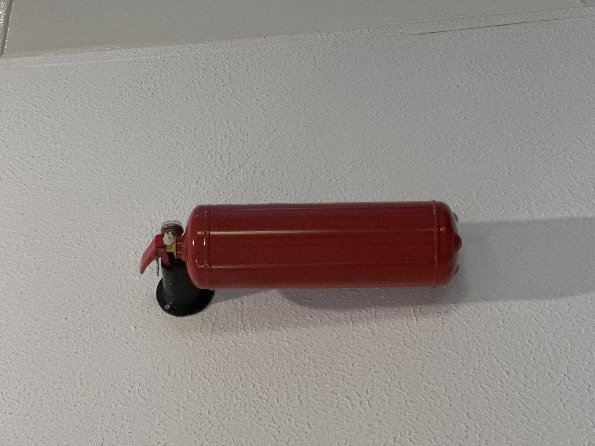 A red fire extinguisher is mounted horizontally on a textured white wall. The extinguisher is secured in a black, circular mount. The wall appears to be indoors, and the lighting is somewhat diffused, creating a soft shadow on the right side of the extinguisher. The exact location in Yerevan, Armenia is not apparent from the image.