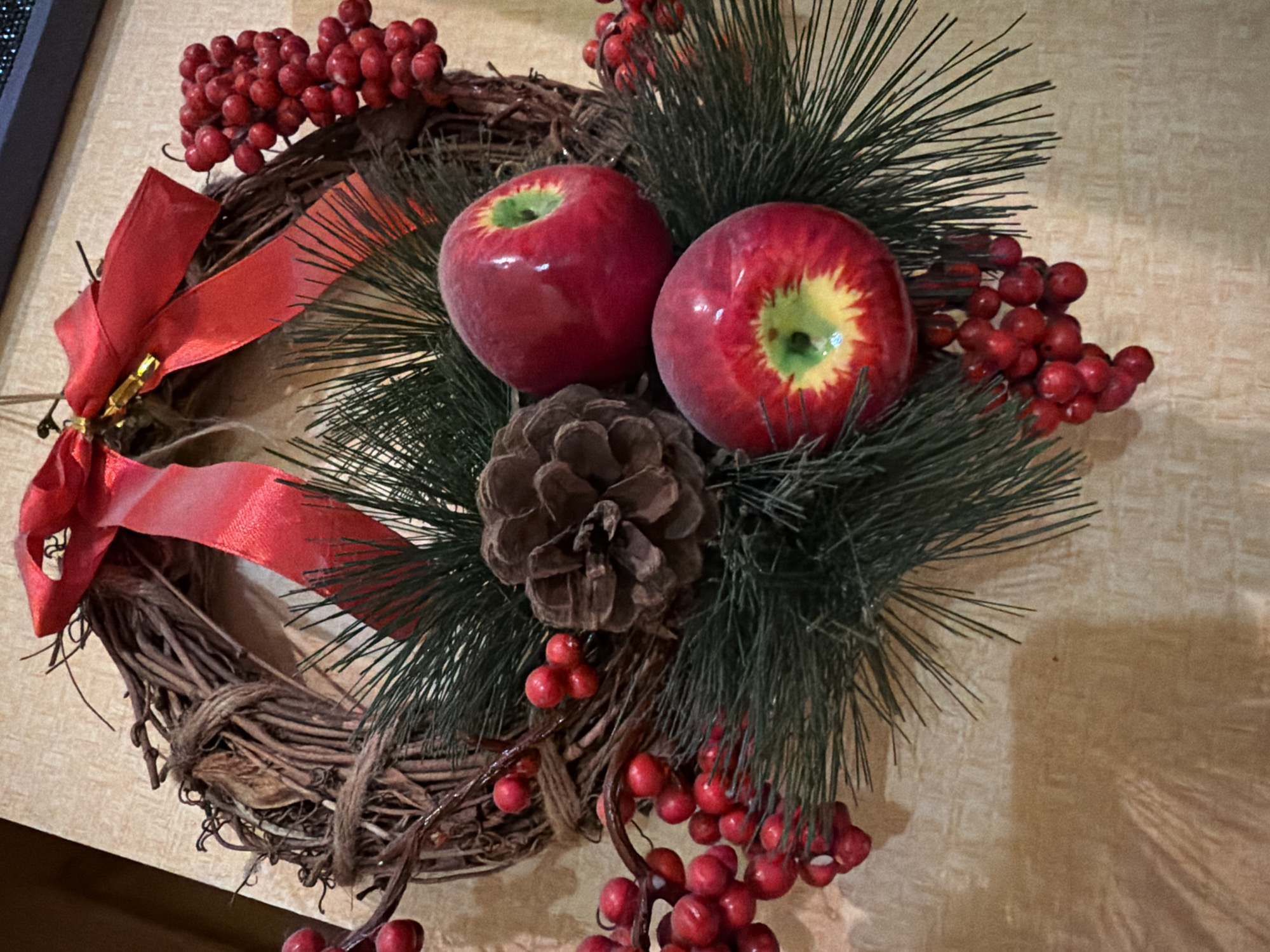 This is a close-up, overhead shot of a rustic Christmas wreath hanging against a textured, beige wall. The wreath is made of intertwined brown branches and decorated with a large red bow at the top, two bright red artificial apples, a pine cone, and clusters of small red berries, interspersed with dark green artificial pine needles. The lighting creates some shadows, suggesting an indoor setting. There are no people or animals visible in the image. The scene is static and appears to be a decorative item. The location in Kyiv, Ukraine is not discernible from the image itself.