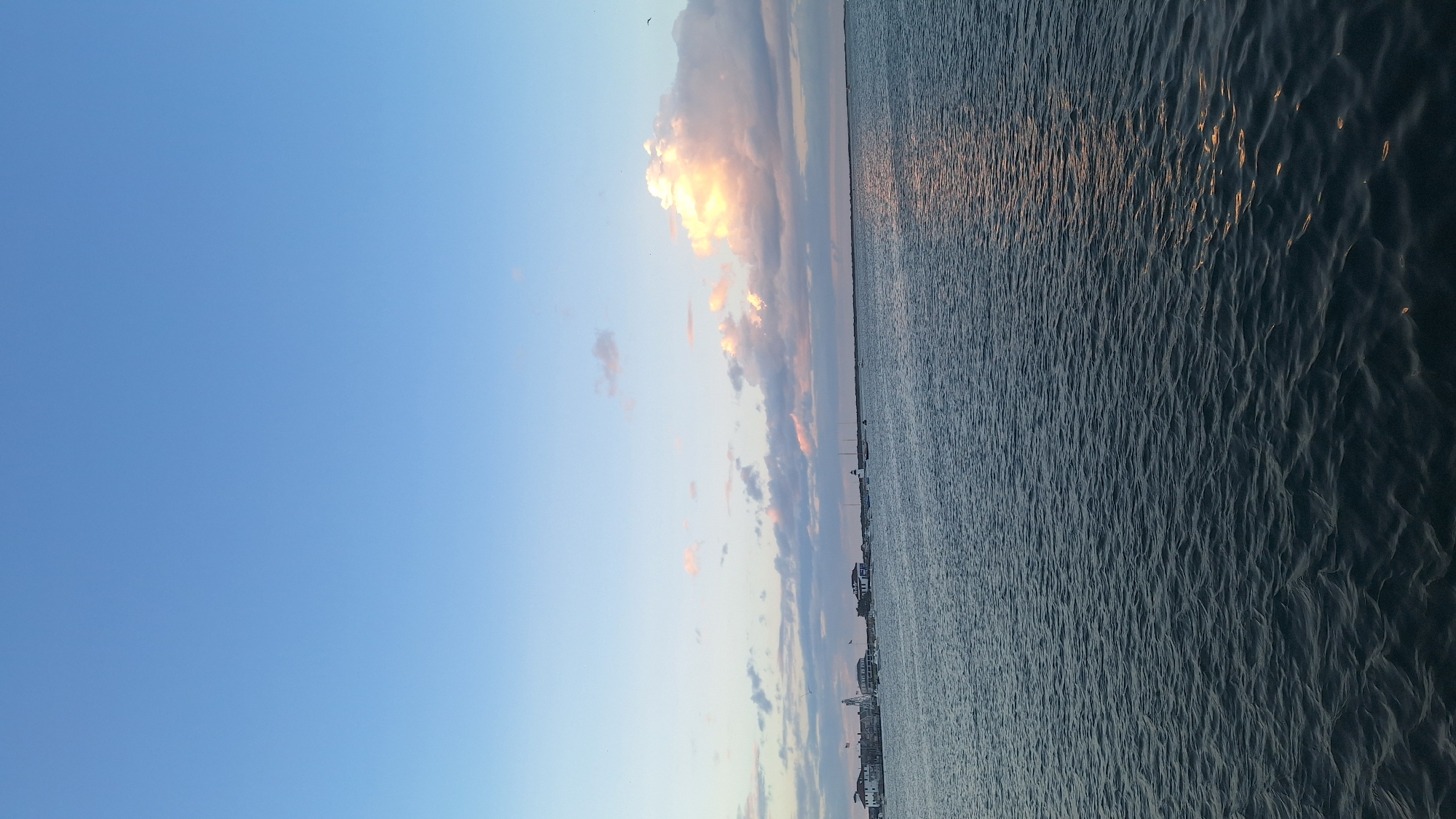 This image depicts a serene body of water with a distant shoreline in Nantucket, United States of America. The sky is a gradient of pale blue, transitioning to lighter shades near the horizon, with a few scattered clouds tinged with pink and orange, suggesting either dawn or dusk. The water in the foreground is dark and rippled, reflecting the soft light from the sky.
The shoreline features a cluster of buildings, appearing to be a small town or village. Among these structures, a white lighthouse is clearly visible. Several sailboats and other smaller vessels are moored or sailing in the water closer to the shore. The overall atmosphere is calm and peaceful, with the natural elements of water and sky dominating the scene, punctuated by the man-made structures of the coastal settlement.