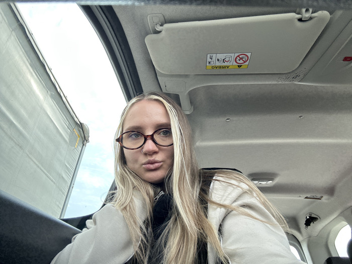 A female subject is shown inside the passenger compartment of a vehicle, in Torbalı, Turkey. The subject, with long, straight blonde hair exhibiting darker roots, wears dark-rimmed oval glasses and a nose ring on her left nostril. She is dressed in a light-colored hooded sweatshirt over a dark undergarment, looking directly at the camera with a pouting expression. Her right arm is positioned on a dark surface, likely a door panel or console. The vehicle's interior features a light-colored headliner and a driver's side sun visor displaying an "AIRBAG" warning label with child seat diagrams. Through the window on the left, a large, light grey canvas tarp or cover, consistent with a truck trailer or industrial structure, is visible against an overcast sky.