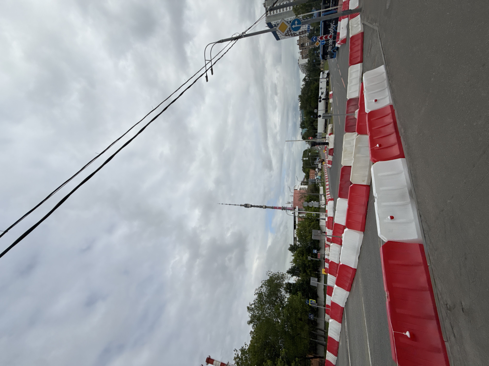 This media file captures an urban street scene in Moscow, Russia, under an overcast sky. The image is oriented vertically but depicts a horizontal landscape.
In the foreground, an asphalt road runs along the right side, bordered by a long, curving line of red and white alternating plastic road barriers. These temporary barriers suggest a designated lane or a special event setup.
The mid-ground features a street lined with trees and a utility pole from which power lines extend across the upper part of the frame. Several street signs are visible, including blue circular signs with white arrows indicating mandatory directions, a yellow diamond sign, and pedestrian crossing signs. Further down the street, various vehicles, including a large white truck and a blue bus, are visible.
In the background, multi-story buildings line the street. Dominating the distant skyline on the right is the very tall, slender Ostankino Tower, a prominent landmark in Moscow, with its characteristic antenna-like top.
The sky is heavily overcast with dense grey clouds, suggesting a cloudy or potentially rainy day.
Visible text includes "Это Электробус." (Eto Elektrobus. - This is an electric bus.) written in Cyrillic on the side of the blue bus. Other visible text is part of standard road signs, such as directional arrows and pedestrian crossing symbols. No people are distinctly visible.