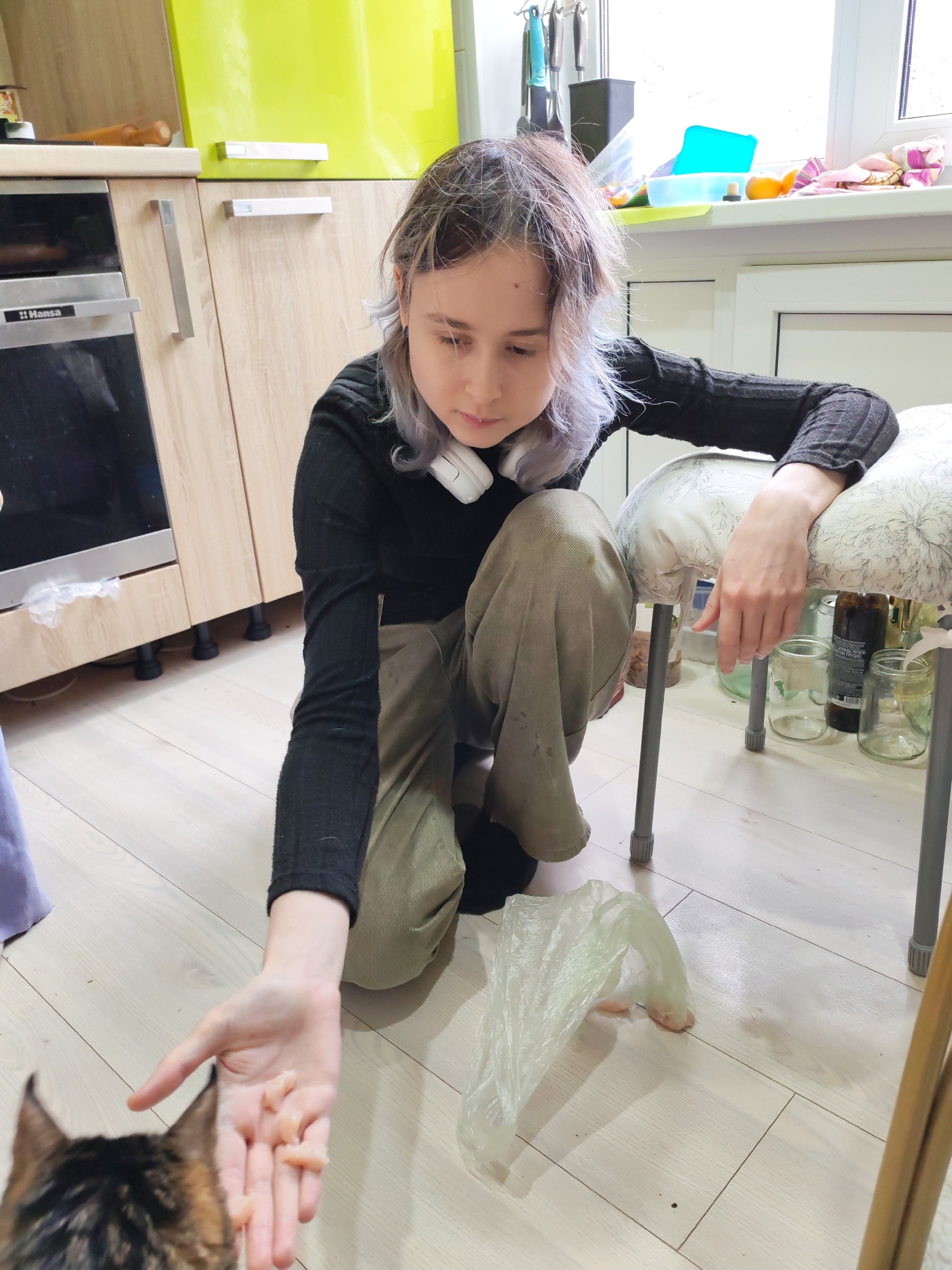 A woman is kneeling on a light wood-toned floor in a kitchen, offering pieces of raw meat from her hand to a cat partially visible in the lower left corner. The woman has light skin, shoulder-length brown hair with grey highlights, and is wearing a black long-sleeved shirt and light brown pants. A pair of white headphones rests around her neck. Behind her is a stool with a patterned cushion. Jars are visible beneath the stool. A plastic bag with additional meat rests on the floor near the woman. In the background are kitchen cabinets in light brown and lime green, along with a black oven and a window above a countertop. The location is the city of Chaikovsky, Russia.