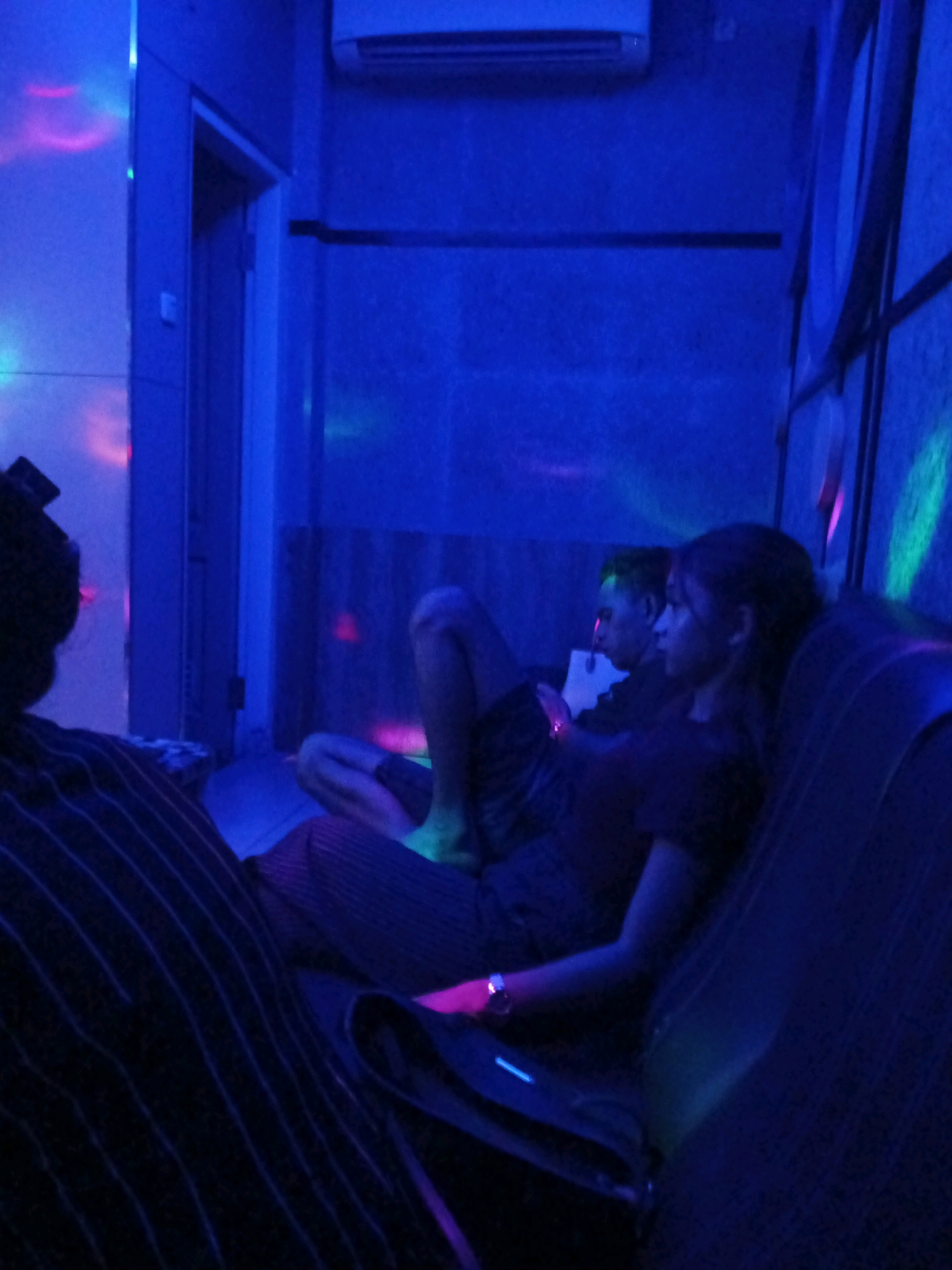 The image depicts a dimly lit room with a blue ambiance, possibly a karaoke lounge or entertainment venue, located in Kalangbret, Indonesia. In the foreground, on the left, a person is seated, facing away from the camera and towards the right. They are wearing a dark shirt with thin vertical stripes. To their right, two individuals are seated on a couch. The person on the left is a male, facing towards the right, with his head slightly tilted down. The person on the right is a female, looking towards the left. She is wearing a dark top and has a watch on her left wrist. The room is decorated with colorful lights, casting vibrant hues of pink, green, and blue onto the walls and ceiling. An air conditioning unit is visible on the upper wall. A doorway is present on the left side of the image. The overall scene suggests a relaxed atmosphere, possibly during a karaoke session or a social gathering.