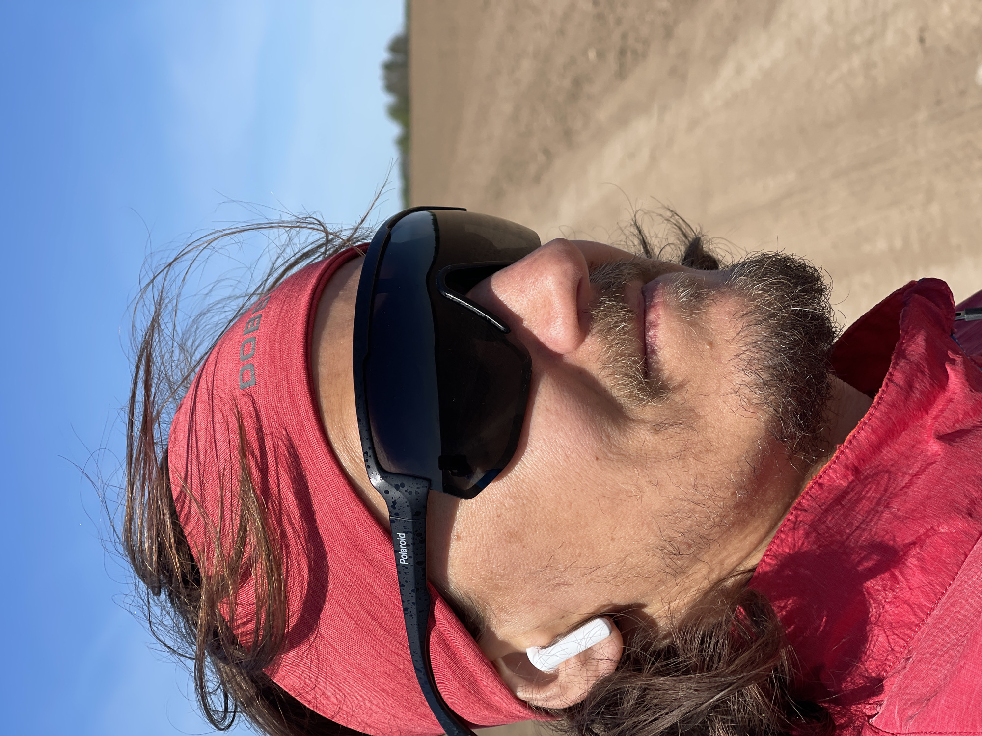 A man is shown in a close-up, angled portrait, wearing sunglasses with the word "Polaroid" visible on the side, a red headband, a red shirt, and a white wireless earbud. He has a beard and long hair. The background consists of a blue sky and what appears to be an agricultural field. While the image suggests an outdoor location, there's no specific visual information tying it to the city of Перевозное, Russia. It is impossible to verify the location based on the image alone.