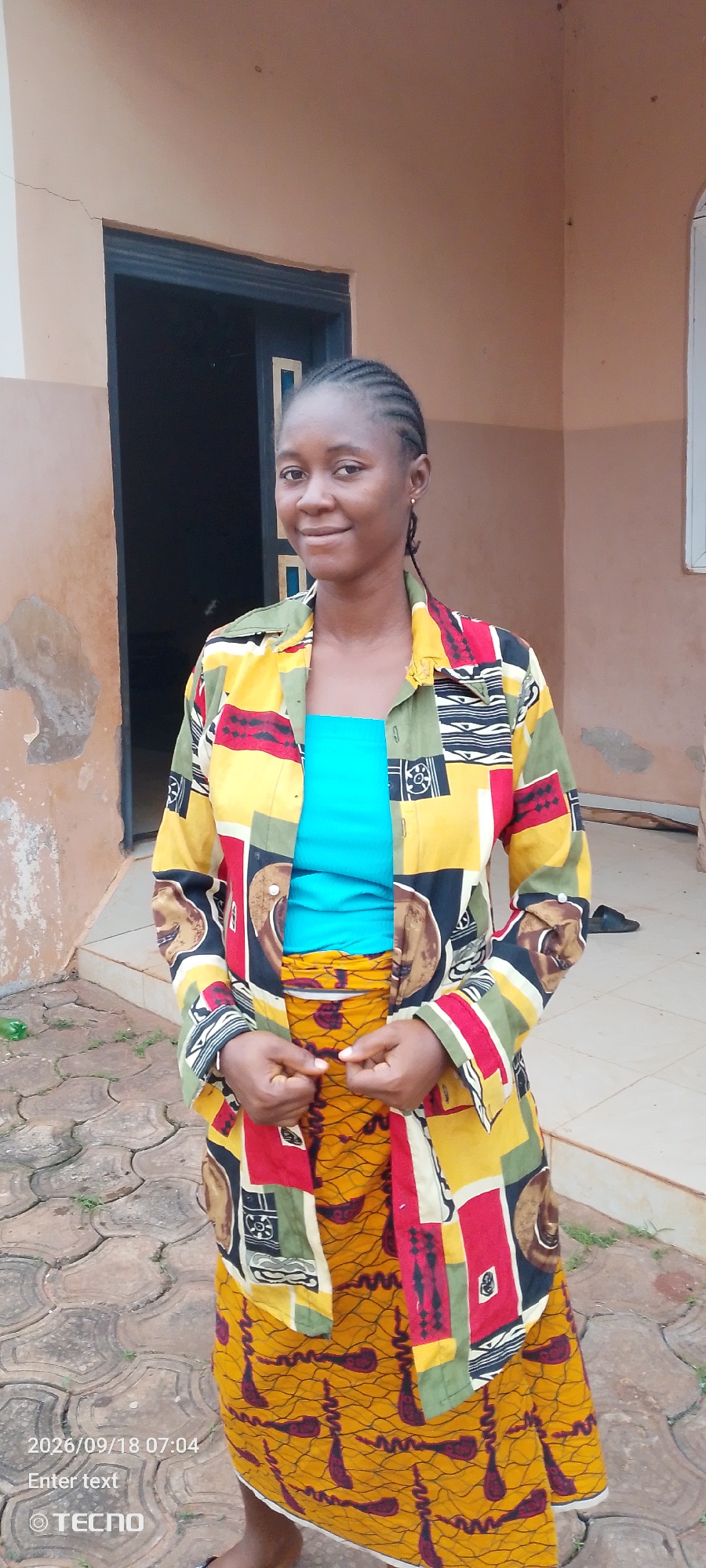 A woman stands outdoors in front of a building. She is wearing a colorful, patterned jacket over a bright blue top, and a matching patterned skirt. The woman has braided hair and is smiling at the camera. The building behind her appears to be made of a light-colored material, with a dark doorway. The ground in front of the building is paved with interlocking stones. The timestamp "2026/09/18 07:04" and the logo "TECNO" are visible in the bottom left corner of the image.