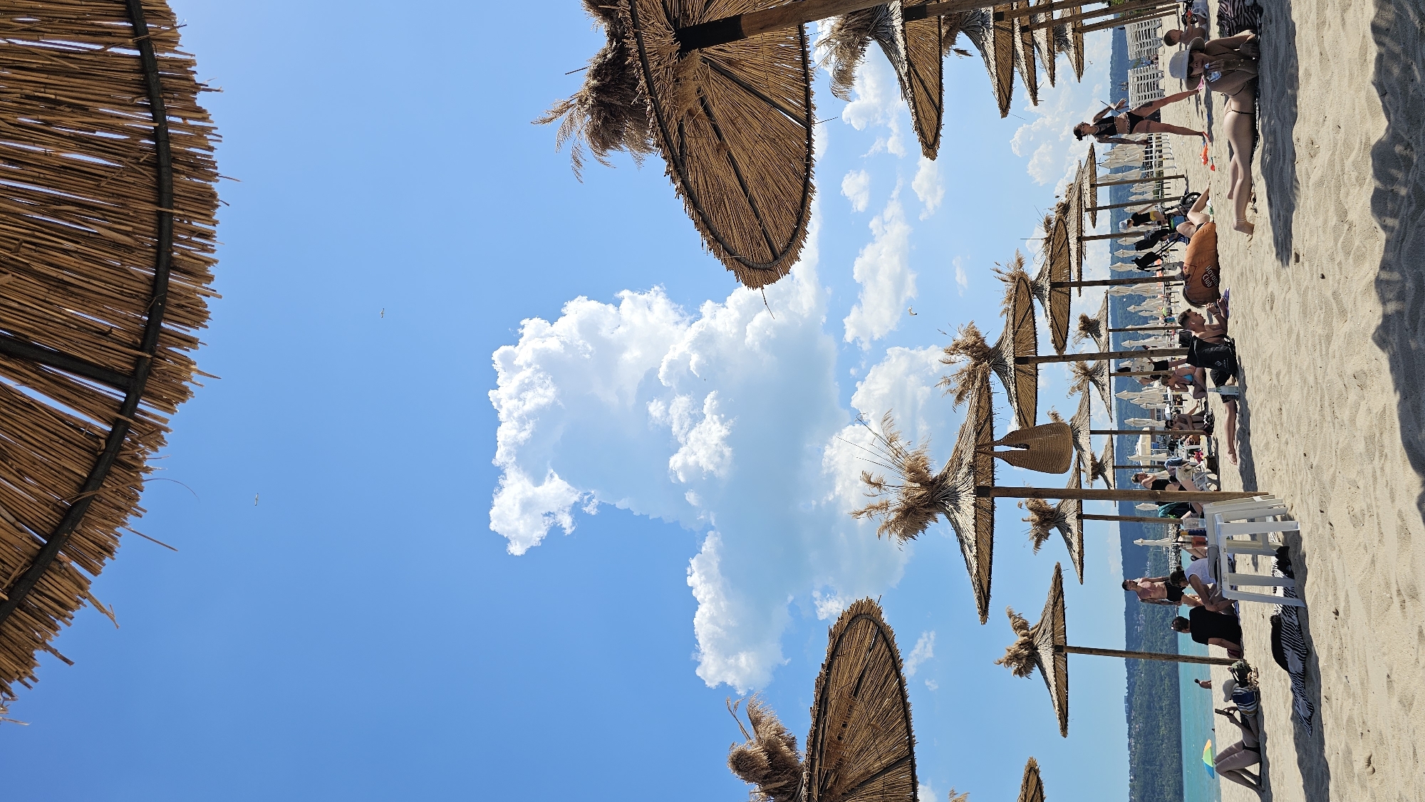 The image depicts a beach scene in Varna, Bulgaria. The perspective is tilted. There is a line of thatched umbrellas extending along the beach. People are lounging and sitting under the umbrellas, and there are beach chairs present. The sea can be seen in the background. Above, the sky is blue with scattered white clouds.