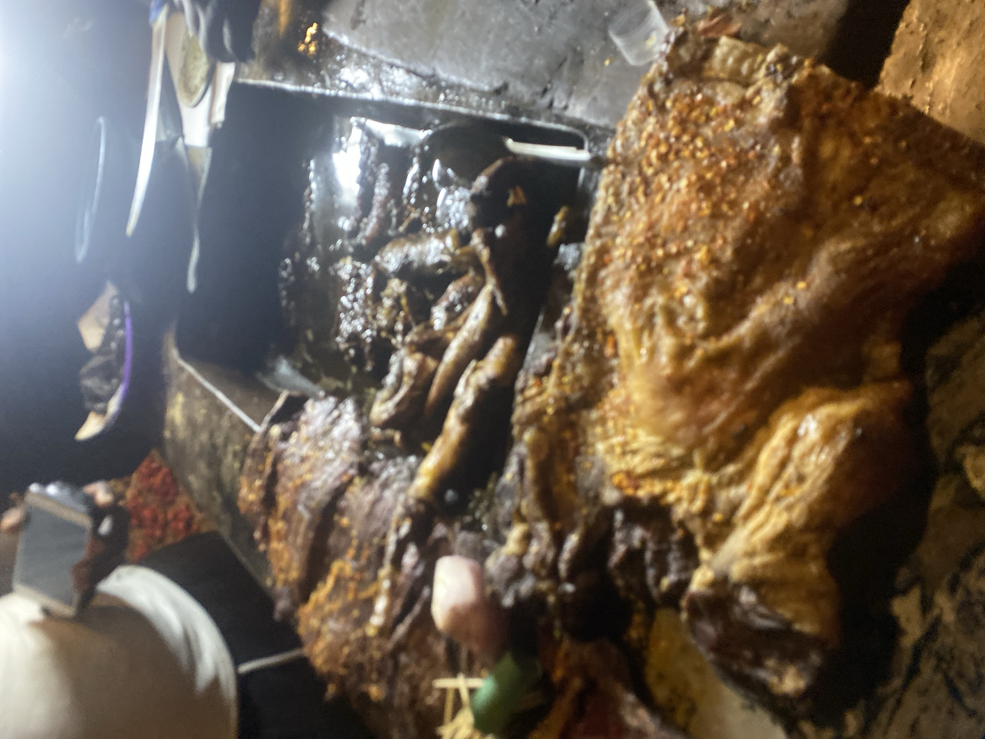 This image depicts a bustling street food scene, likely at night in Bukuru, Nigeria, centered around the preparation of a large quantity of roasted or grilled meat. The primary subject is a substantial piece of meat, possibly beef or goat, generously seasoned with what appears to be red pepper flakes and other dry spices, resulting in a golden-brown, somewhat crispy texture. It rests on a large, well-used, dark metal cooking surface or grill.
To the left of the main piece of meat, smaller, darker cuts of meat or offal are visible, possibly stewing or simmering in a liquid within another section of the cooking apparatus. The scene is illuminated by bright, artificial light sources, casting reflections on the greasy surfaces and highlighting the rich textures of the food.
In the lower-left foreground, the top of a person's head and shoulder are visible, along with their hand holding a smartphone, suggesting they are capturing the scene. Another individual's arm, holding a long tool that resembles a knife or spatula, can be faintly seen in the upper-left, indicating active cooking or serving. Small pieces of green and pinkish ingredients, possibly lime or onion, are scattered near the seasoned meat, likely intended as accompaniments. The overall setting appears to be a rustic outdoor or semi-outdoor food stall, indicative of a vibrant local food culture. There is no visible text in the image.