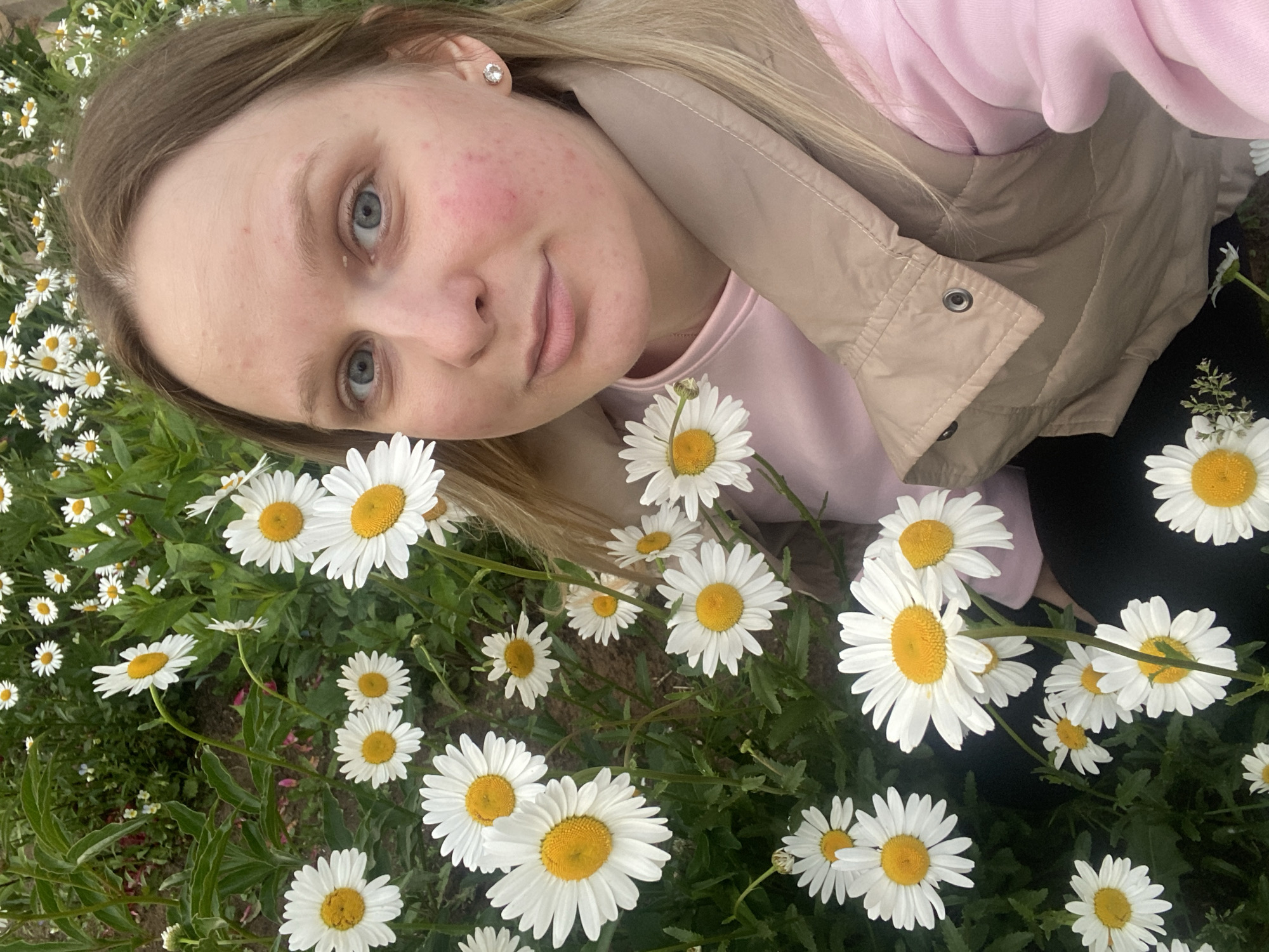 A young female with long, light brown hair and blue eyes is captured in a close-up, selfie-style photograph amidst a dense cluster of white daisies. Her head and upper torso occupy the right side of the frame, angled slightly to her left. She presents a neutral expression with slightly parted lips, looking directly at the camera. Her pale skin shows areas of redness on her cheeks, and a silver stud earring is visible in her right ear. She wears a light pink long-sleeved top beneath a beige quilted vest. The numerous white daisies, characterized by their bright yellow centers and green foliage, densely populate the foreground and background, with glimpses of dark soil between the plants, indicating an outdoor, natural setting.