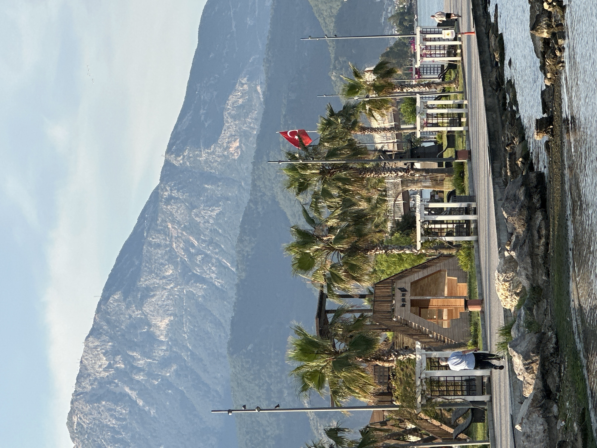 The image displays a scenic coastal view in Fethiye, Turkey, under a bright, clear sky. Towering mountains, likely part of the Taurus range, dominate the background, their craggy, tree-covered slopes rising steeply.
In the mid-ground, a paved promenade stretches along the water's edge, lined with numerous tall palm trees. Interspersed among the palm trees are white-framed structures, possibly seating areas or small kiosks, and streetlights. A prominent red Turkish flag, featuring a white star and crescent, flies from a pole, signaling the country. Behind the palm trees, a few small, distinctively designed wooden buildings with gabled roofs are visible.
Several people are present on the promenade. One individual, wearing a light shirt and dark trousers, is seen walking away from the viewer. Other figures are discernible further down the path, near the water. To the right, the calm water of a bay or sea laps against a rocky shoreline, where some green vegetation grows on the rocks.
The sky is a pale blue with scattered wispy clouds. A small flock of birds is visible flying high above the mountains, and other birds are perched on the streetlights along the promenade. The lighting suggests it is daytime, likely late morning or early afternoon, with soft shadows indicating the sun's position. The scene evokes a tranquil and picturesque coastal environment. No visible text is legible on any signs or structures.