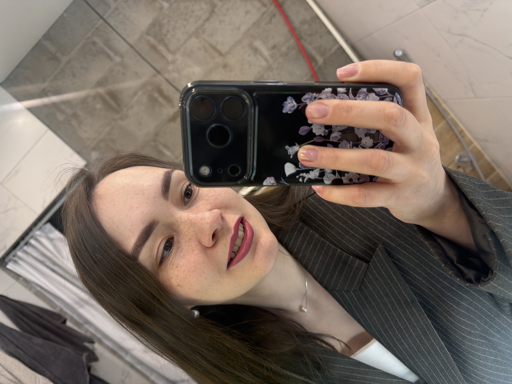 A woman is taking a selfie in what appears to be a bathroom. She is holding a black smartphone with a floral design case up towards the mirror, capturing her image. The woman has light skin, freckles, dark brown hair, and is wearing a gray pinstripe blazer over a white top. She is wearing a silver necklace with a pearl pendant. The background shows a tiled wall, a partially visible red pipe, and what looks like a towel rack with clothing. The image could potentially be located in Chaikovsky, Russia, as requested, but without further information to confirm the location this can't be verified.