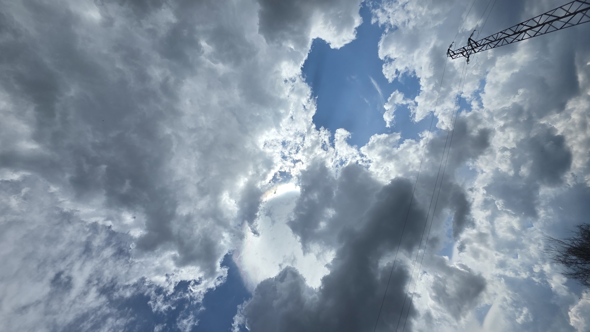 The image captures a dramatic sky filled with a mix of dark and bright clouds, looking upwards towards the zenith. The dominant subjects are the expansive sky and various cloud formations. Large, dark grey cumulus or stratocumulus clouds cover much of the frame, interspersed with patches of bright blue sky. The sun is partly visible behind the clouds, casting intense rays of light and creating a bright, almost ethereal glow.
A notable natural phenomenon is visible: a circumhorizontal arc or iridescent cloud displaying a distinct rainbow-like spectrum of colors (red, orange, green, blue) on the lower edge of a cloud illuminated by the hidden sun, slightly to the left of the center. Beams of sunlight, also known as crepuscular rays, can be seen breaking through the cloud cover.
In the upper right portion of the image, the metal lattice structure of a high-voltage power line tower is prominently visible, extending diagonally across the sky. Associated power lines run downwards from the tower. On the far right edge of the frame, sparse, dark, bare tree branches or shrubbery are partially captured. A tiny dark speck, possibly a bird or insect, is discernible against the darker clouds in some areas.
The scene depicts daytime conditions, likely mid-day to late afternoon, given the intensity of the light breaking through the clouds. The weather appears dynamic, suggesting a transition from or to an overcast state, perhaps after a rain shower or preceding one. The setting is outdoors, likely in a rural or semi-urban environment, with the power lines hinting at human infrastructure. The location context provided is Drenkovo, Bulgaria. There is no visible text in the image.